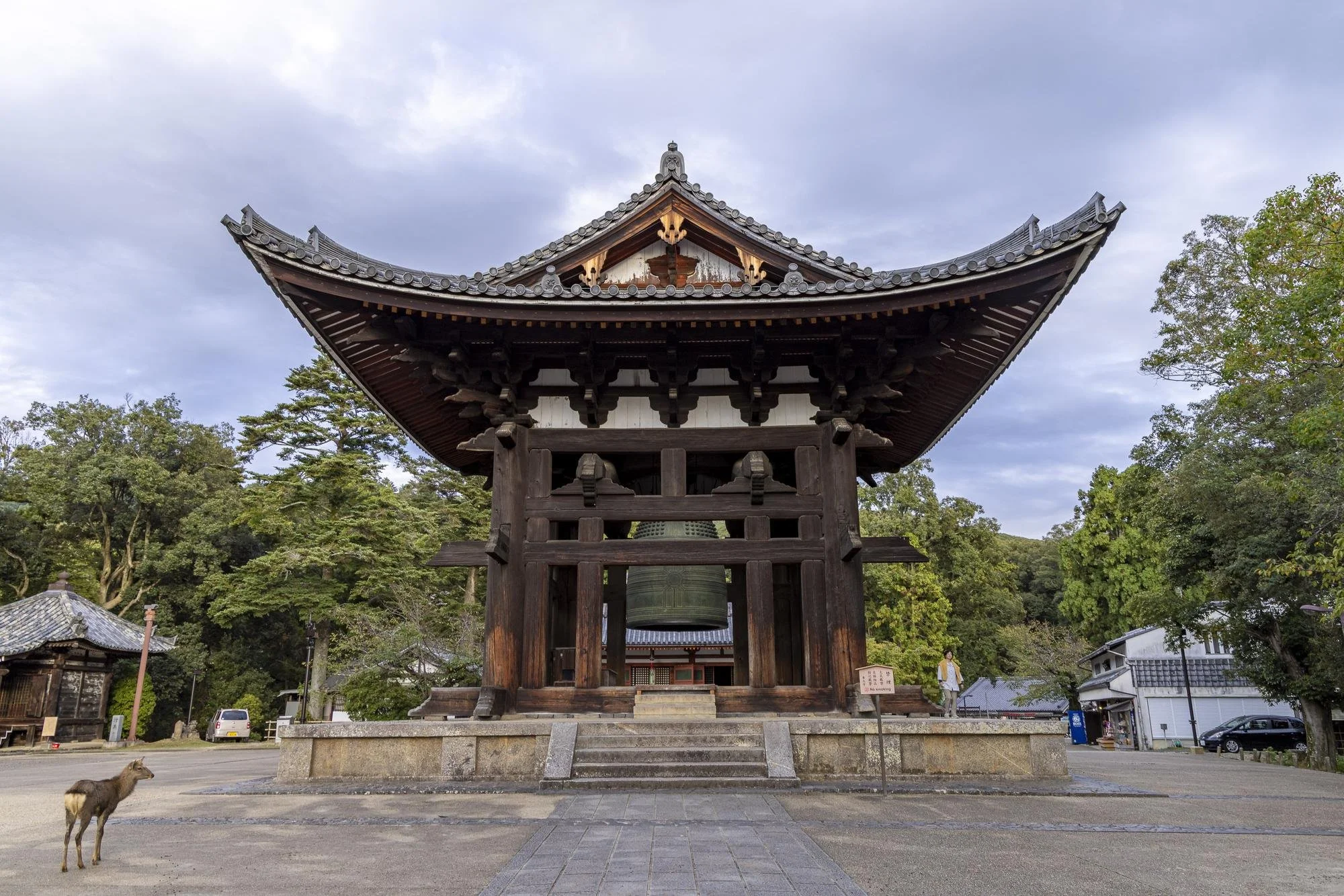 A traditional Japanese temple bell tower with a large bell inside, surrounded by greenery and an overcast sky.