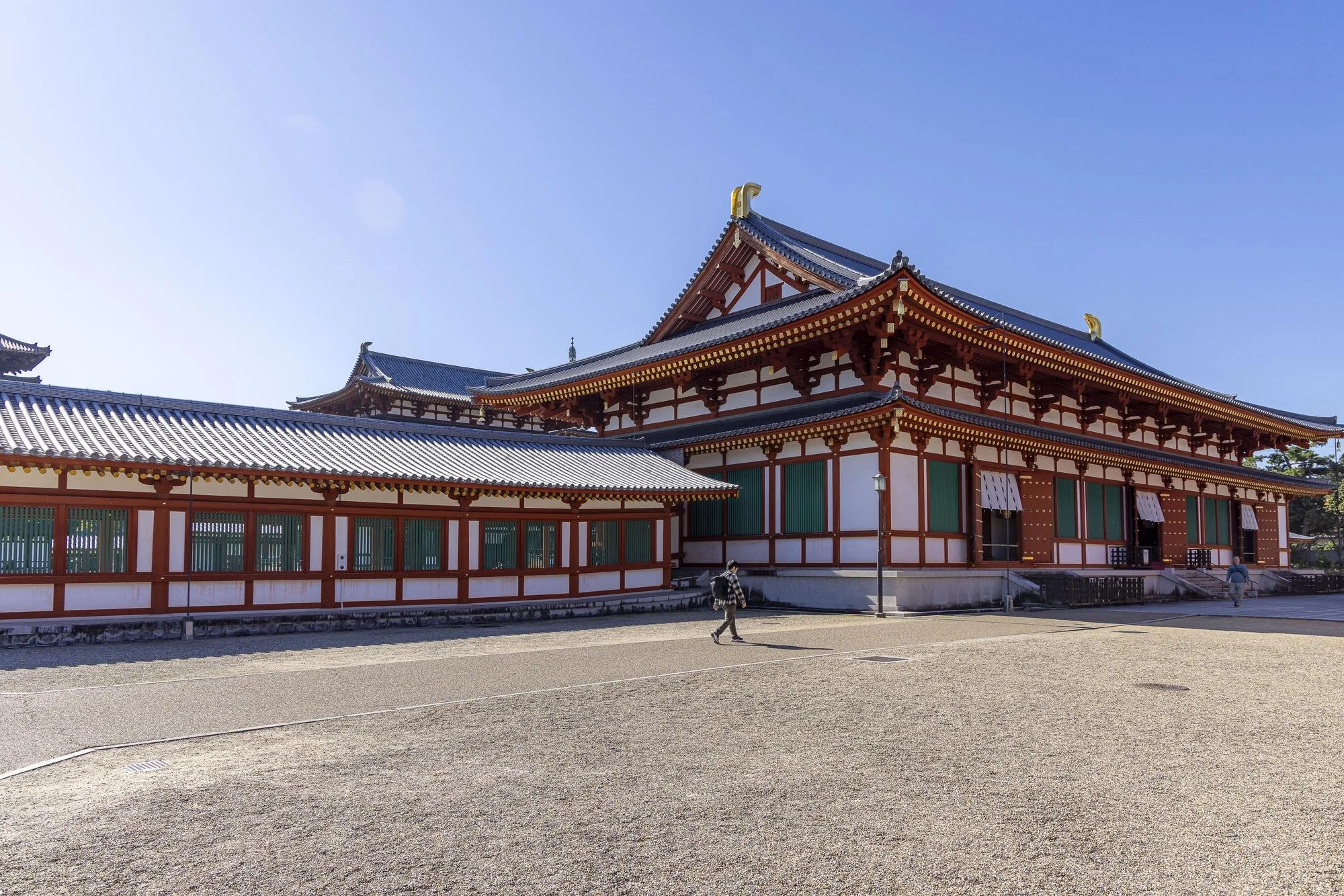 Traditional Japanese temple with red and white walls, dark tiled roof, and decorative golden accents, set against a clear blue sky, with a gravel courtyard and a few visitors walking around.