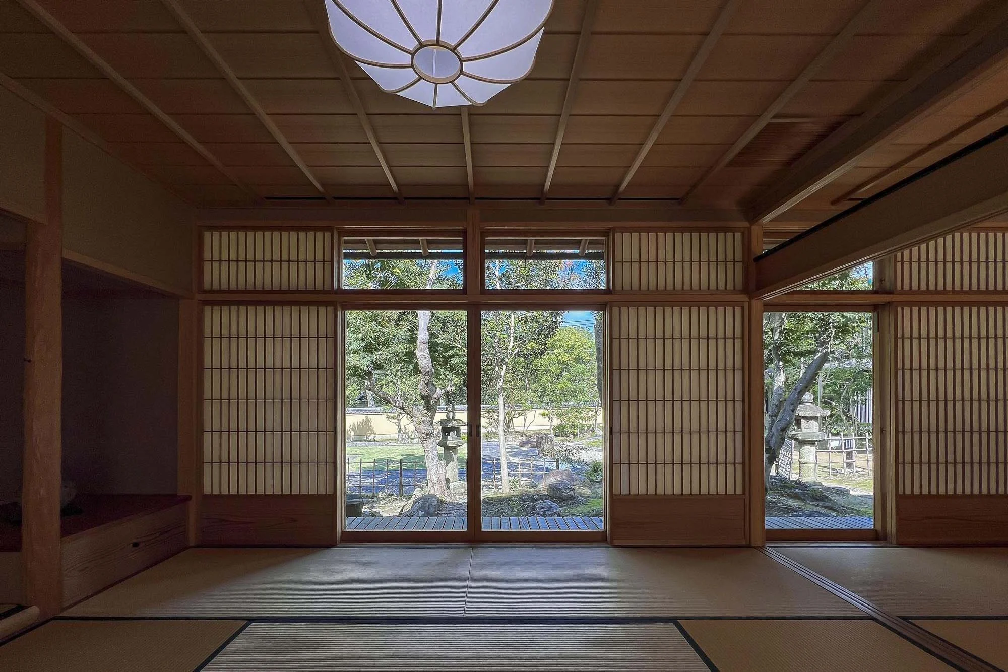 Traditional Japanese room with tatami mats and sliding shoji doors opening to a peaceful garden with trees and rocks.