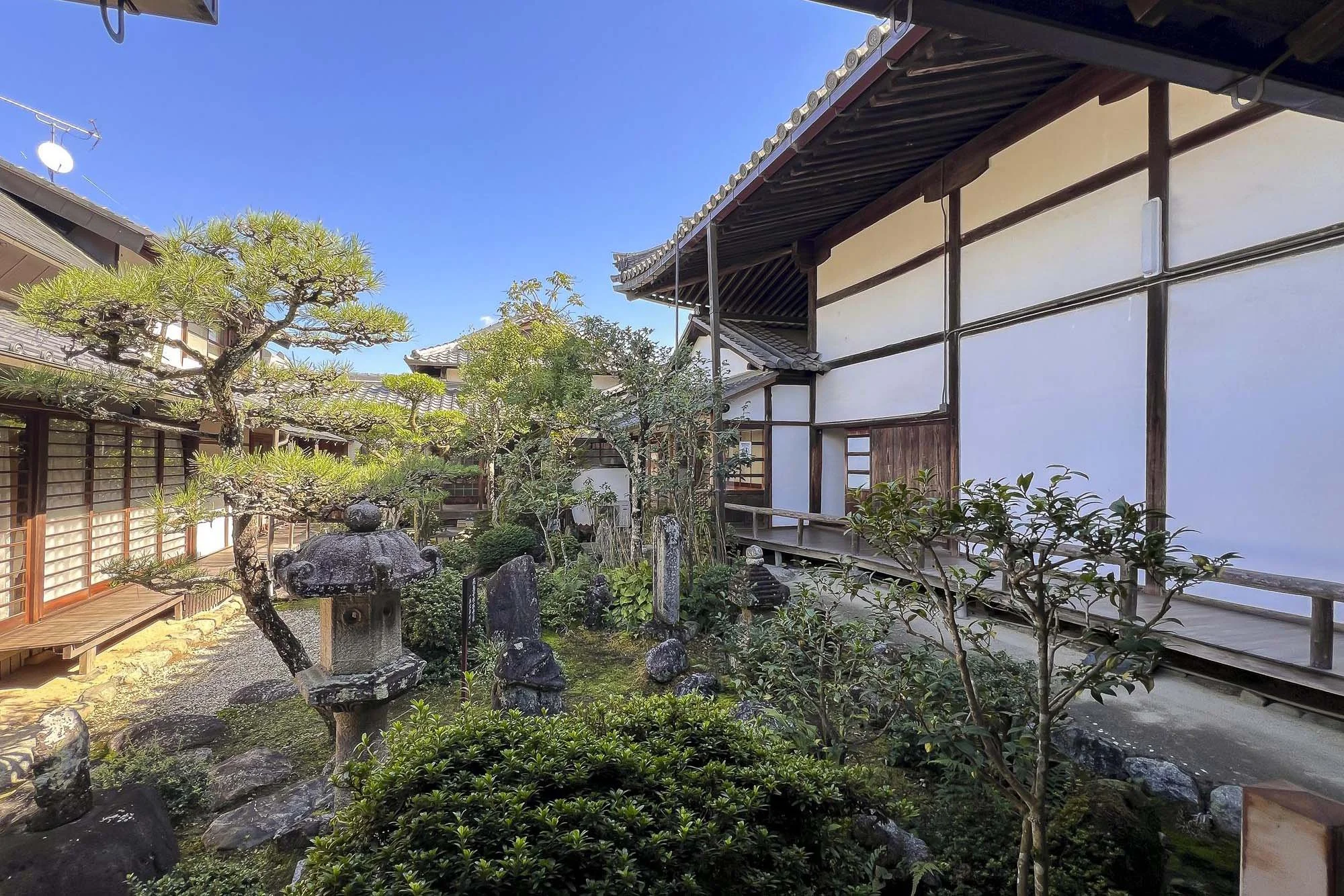 Traditional Japanese garden with rocks, trees, and a stone lantern, adjacent to a building with wooden framing and white walls.