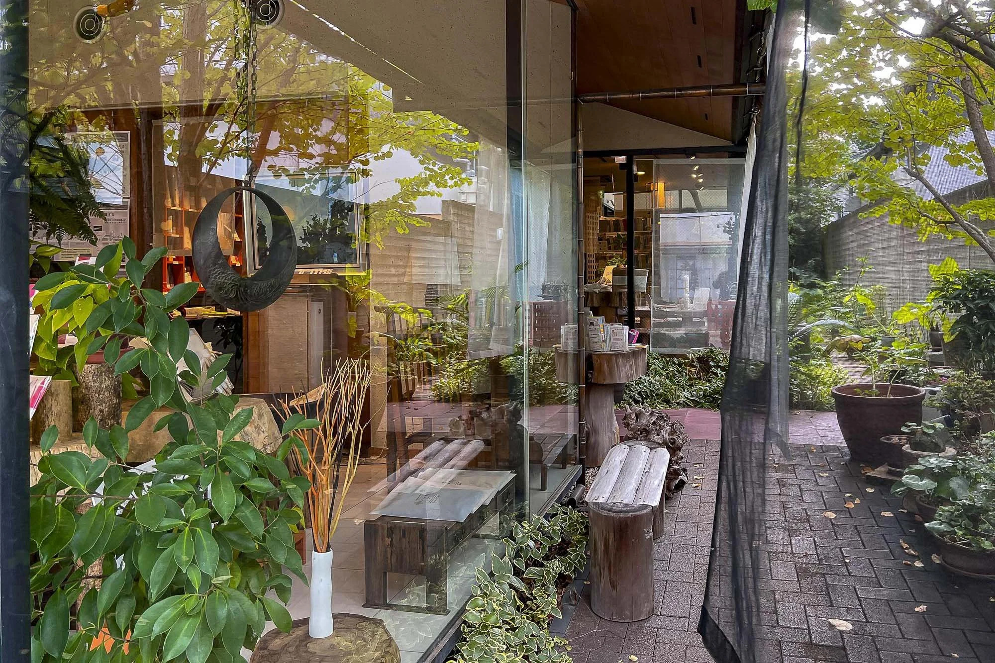 View through glass window of an indoor space with plants, wood furniture, and decorative items, with outdoor plants and brick pathway visible outside.