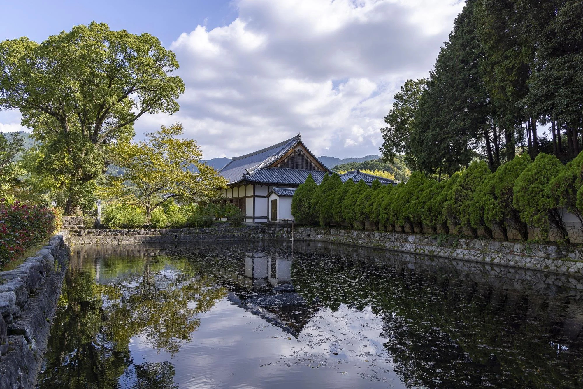 A traditional Japanese building next to a pond with reflections of trees and the building, surrounded by lush greenery and a partly cloudy sky.
