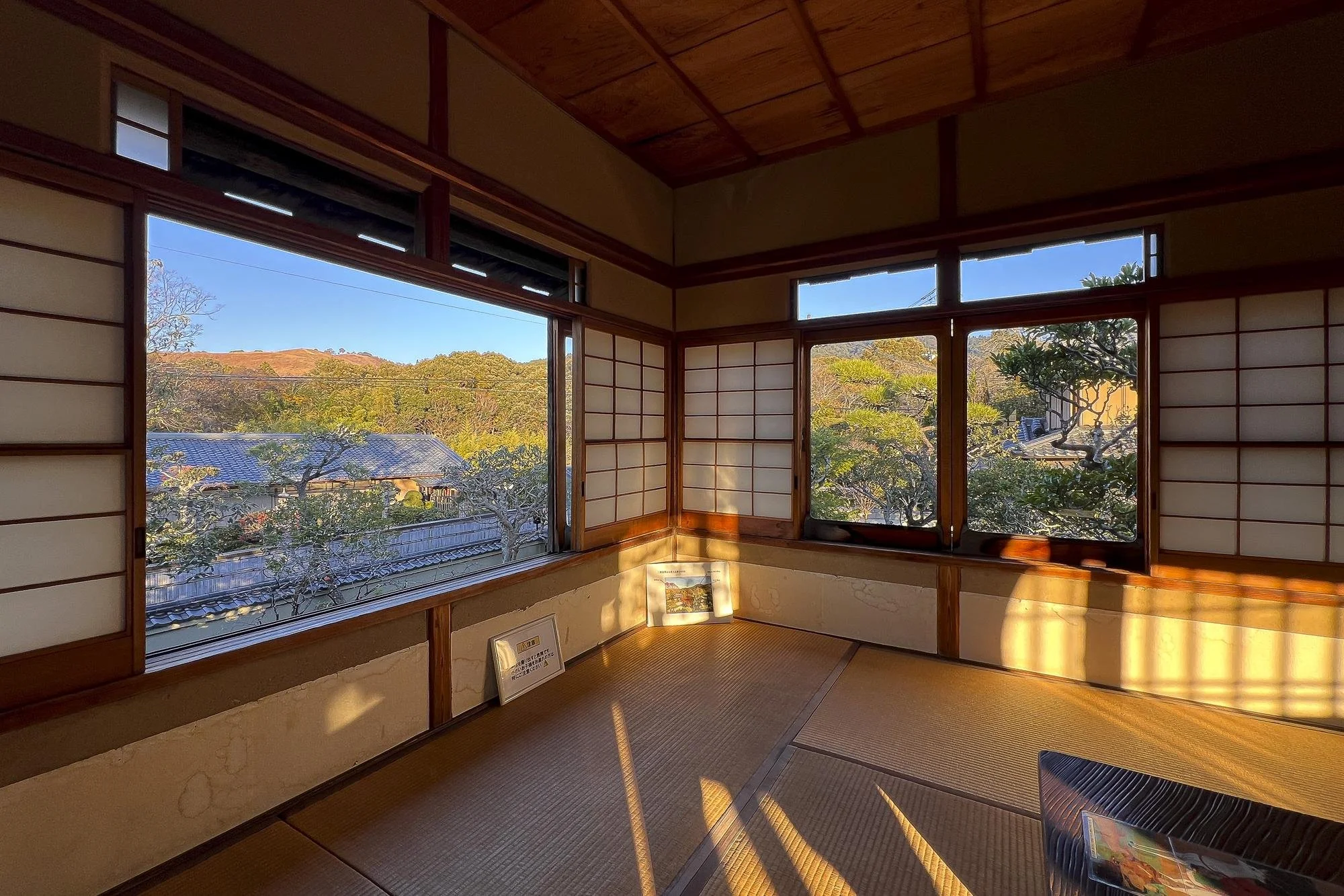Sunlight streams into a traditional Japanese room with tatami mats and shoji screens, revealing a scenic view of trees and mountains outside.
