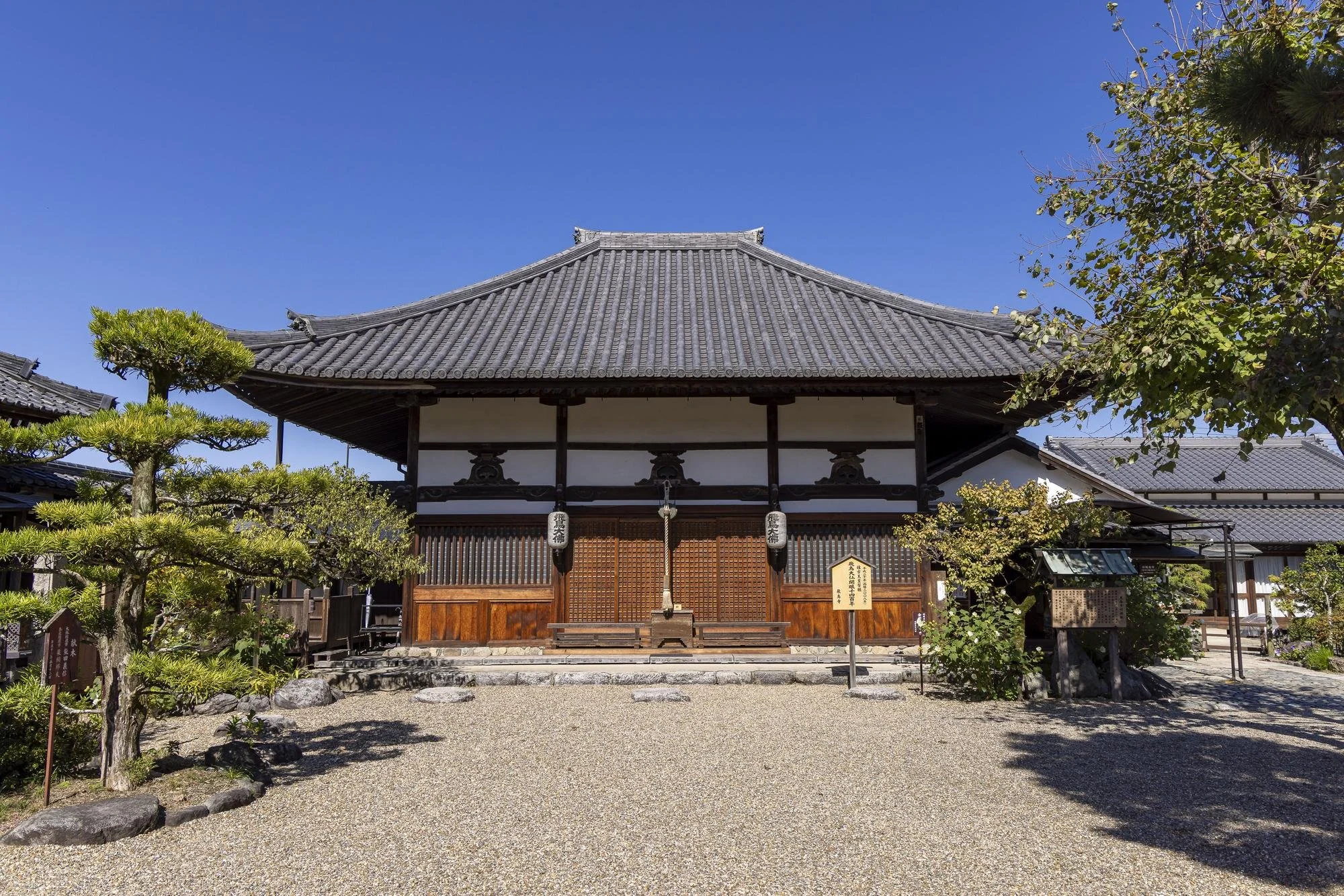 Traditional Japanese temple with wooden doors, decorative lanterns, and a gravel courtyard, surrounded by trees under a clear blue sky.