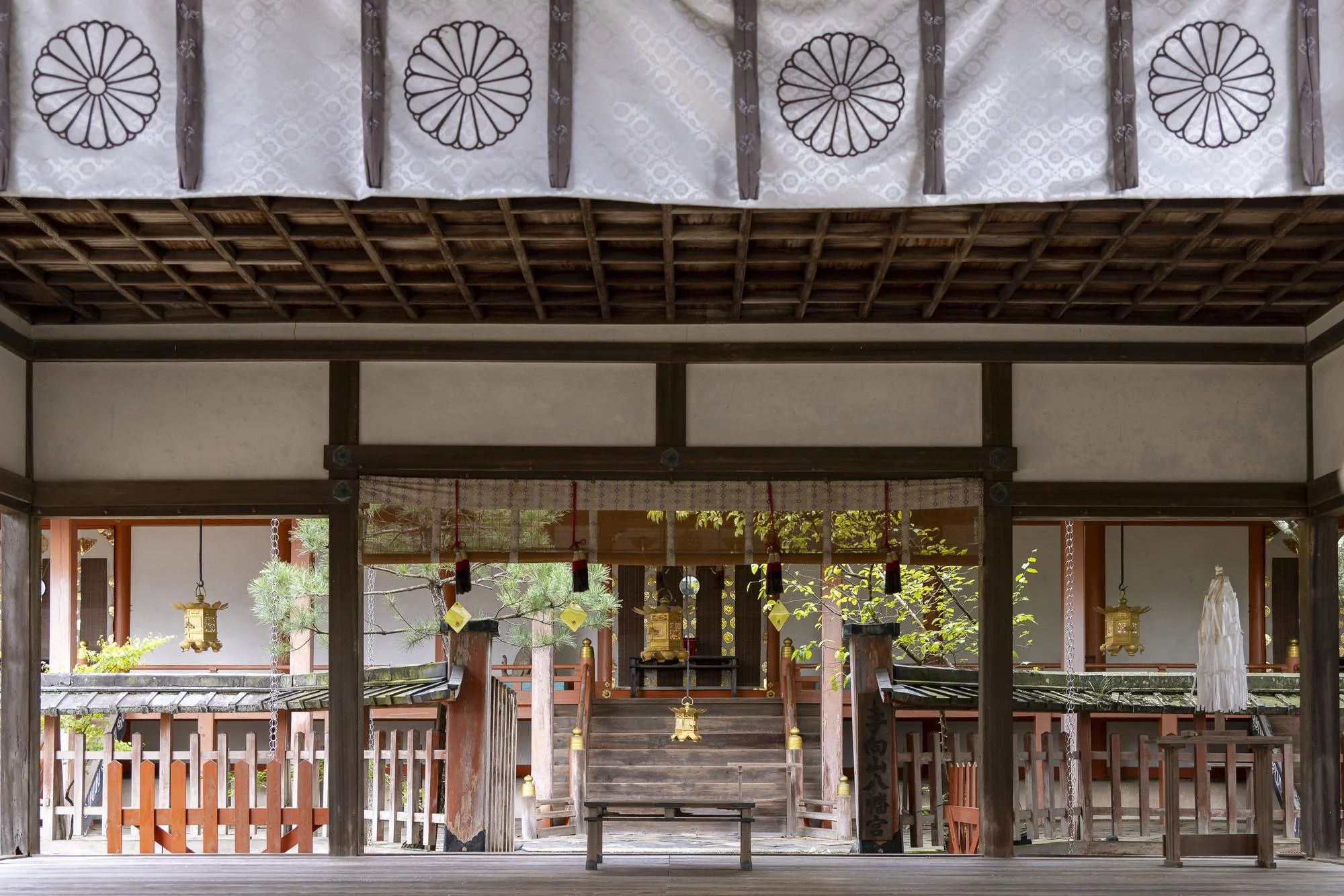 Interior view of a traditional Japanese shrine with wooden steps leading to a sacred space, adorned with hanging lanterns and decorative drapes. Greenery visible in the background.