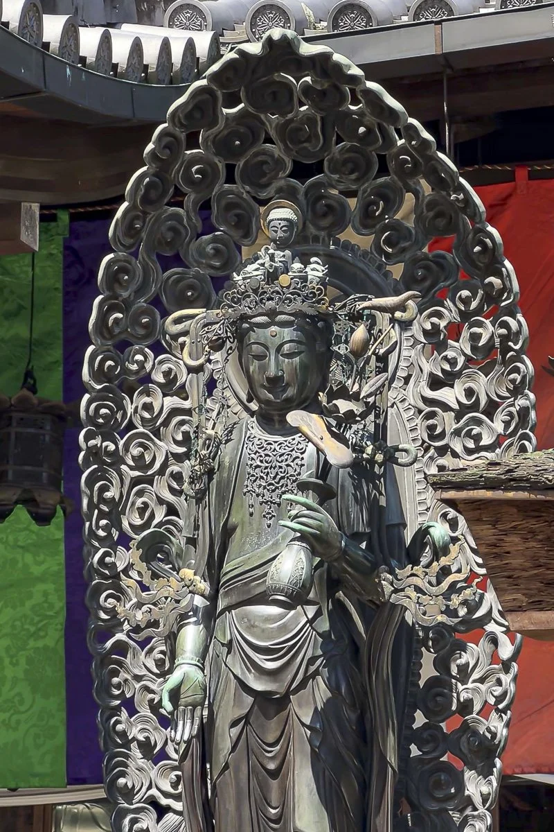 A detailed black wooden statue of a female deity with ornate headgear, surrounded by swirling carved wood, holding a flute and vases, with colorful fabric banners in the background.