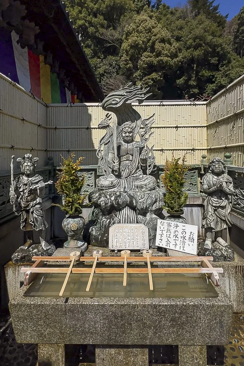 A traditional Japanese altar with statues of warriors, one holding a sword and another holding a book, in front of a bamboo fence and trees. There are small potted trees, a water basin with water ladles, and Japanese signs.