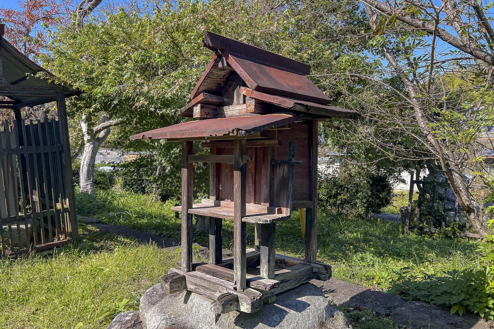 A small wooden shrine on a rock, with a roof, in a grassy yard with trees and a fence in the background.