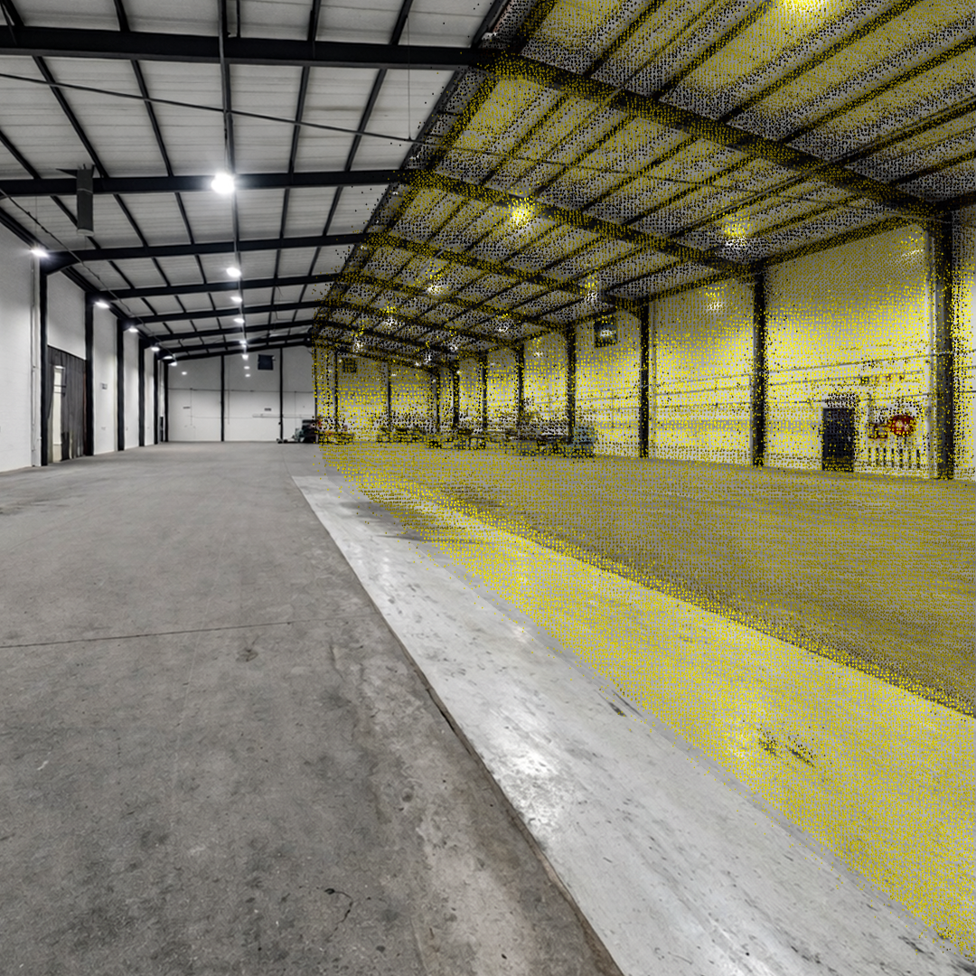Empty indoor warehouse or large garage with a concrete floor, metal beams, and high ceiling. Bright overhead lights illuminate the space. Walls are plain, with some equipment and tools visible on the right side.