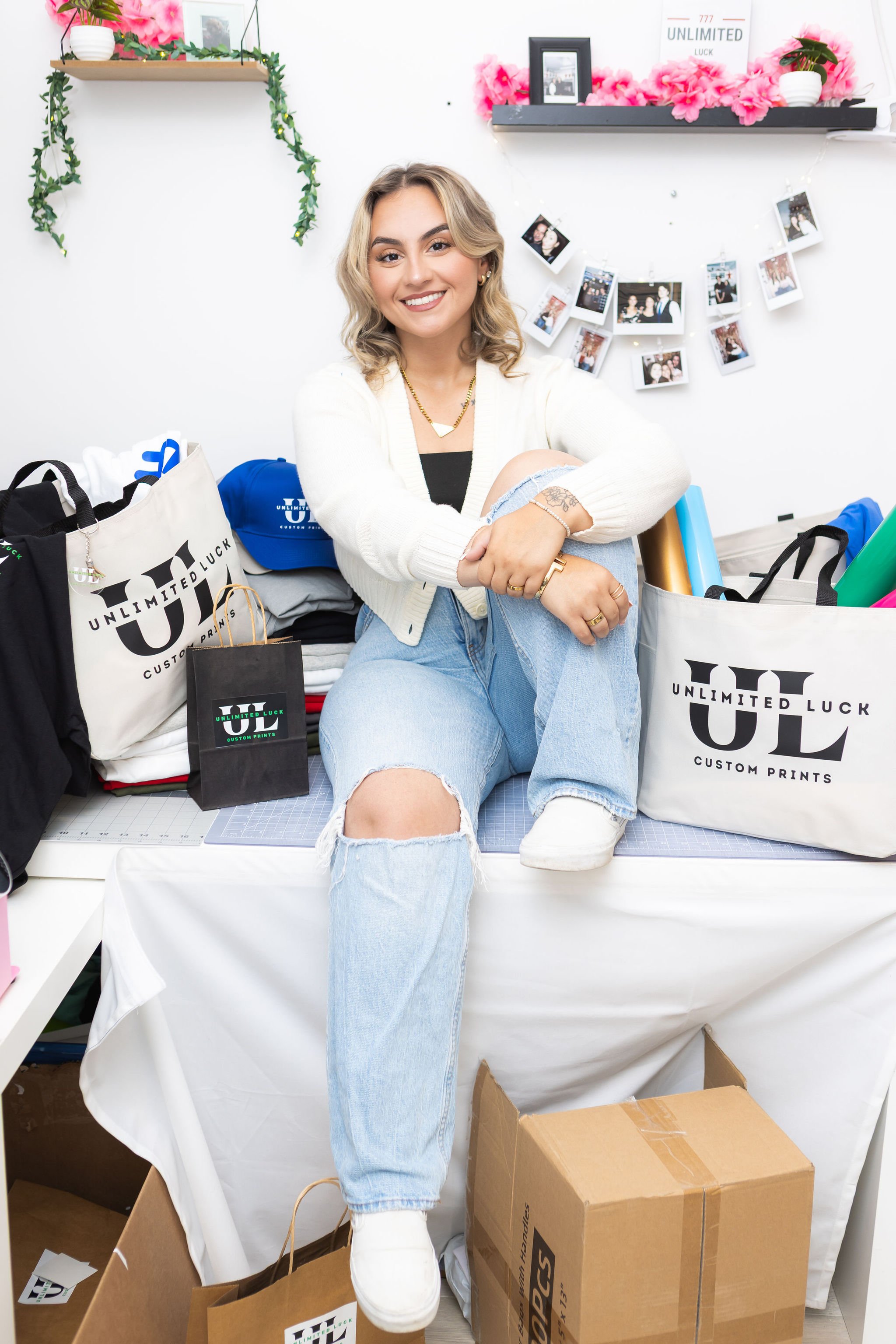 A young woman with blonde hair and light skin sitting on a table surrounded by shopping bags, cardboard boxes, and gift-wrapped items. She is smiling, wearing a white sweater, ripped jeans, and white sneakers. The background has a white wall with photos, pink flowers, and decorative shelves.