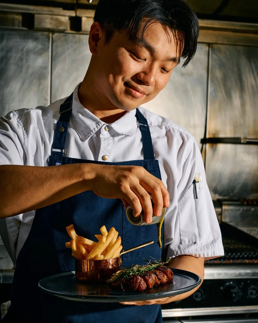 Chef garnishing a plated meal with fries and a small copper cup on a dark plate.