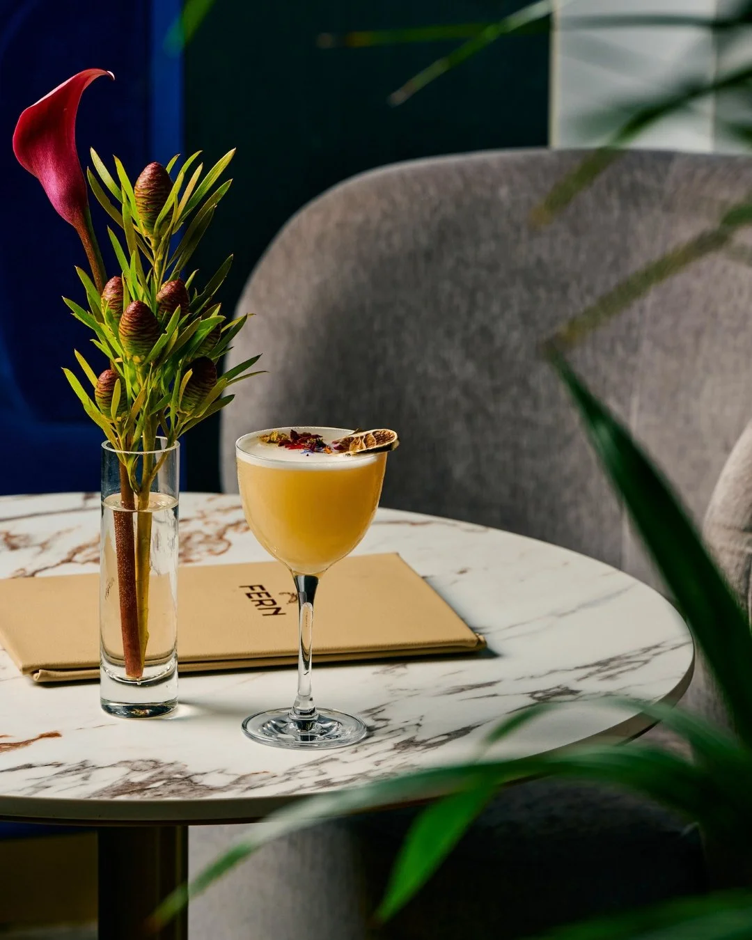 A marble tabletop with a yellow cocktail garnished with dried fruit and herbs, a tall vase with pink calla lily and green foliage, a beige menu, and beige chairs in a stylish setting.
