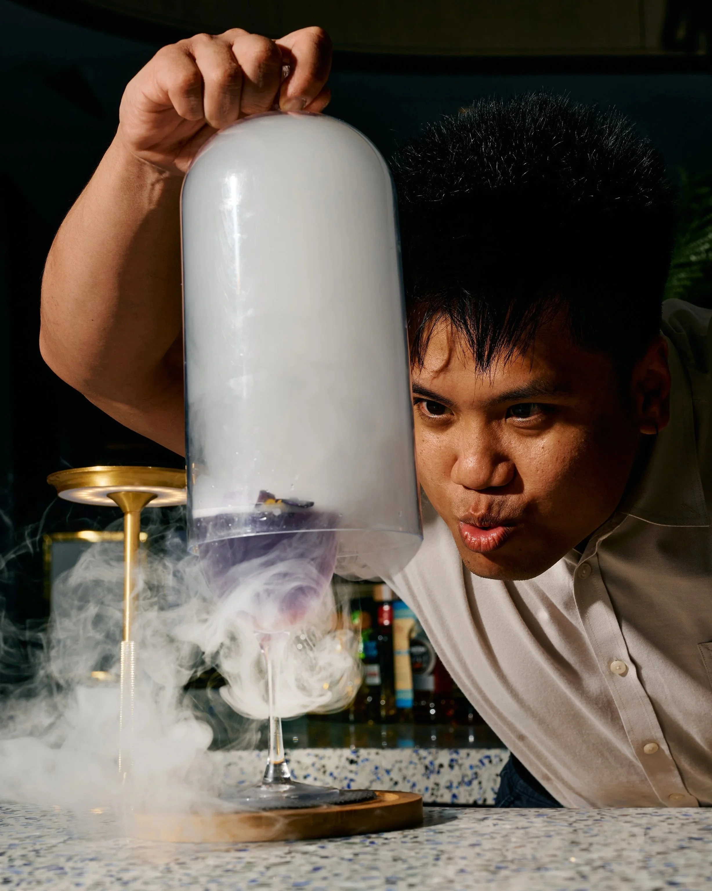 A man in a white shirt is leaning over a table, with a tall glass containing a white smoke effect and a fireworks-like display in the background.