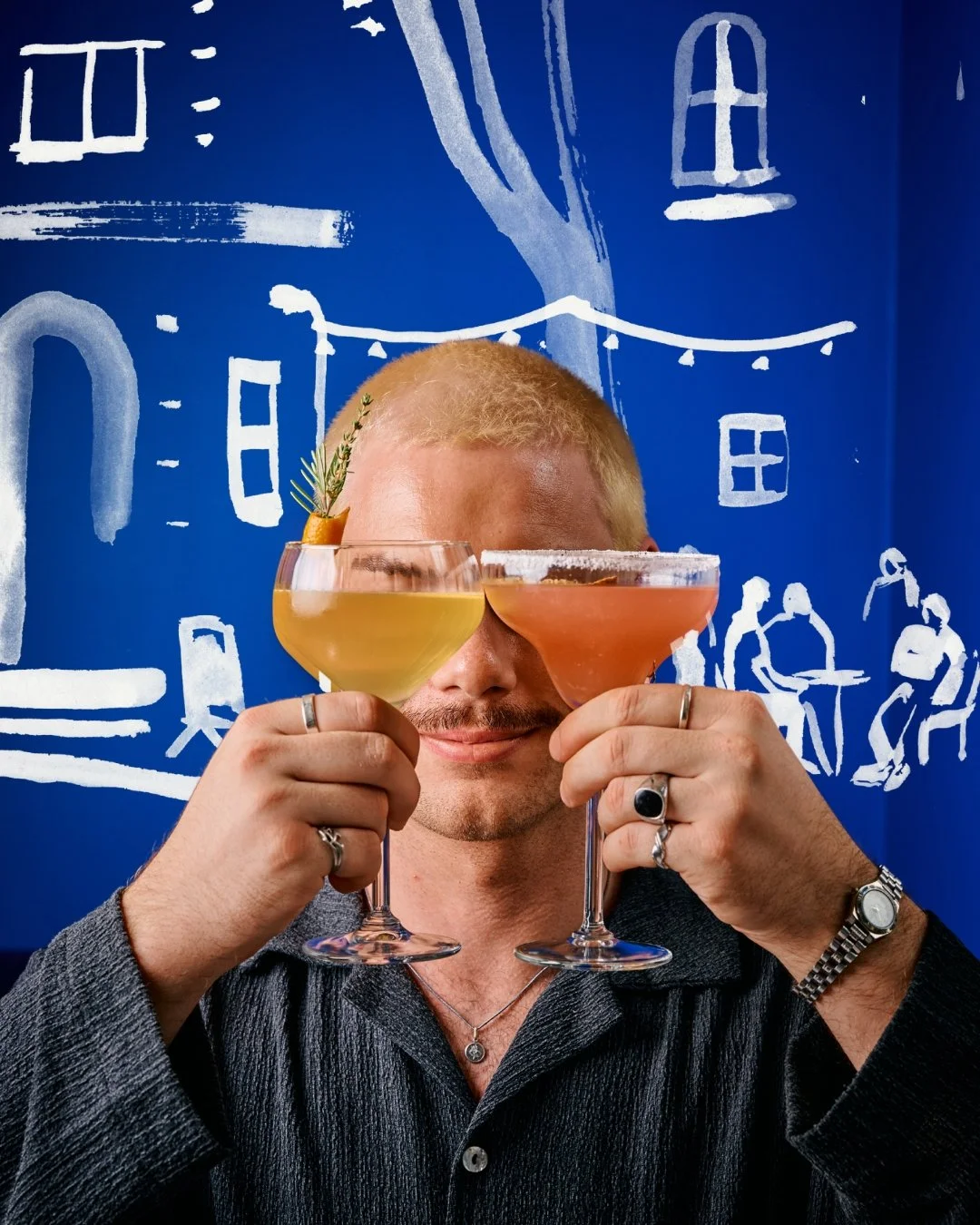 A man with blond hair and rings on his fingers holding two cocktails, one yellow with a garnish and one pink, in front of a blue wall with white line drawings of buildings and people.