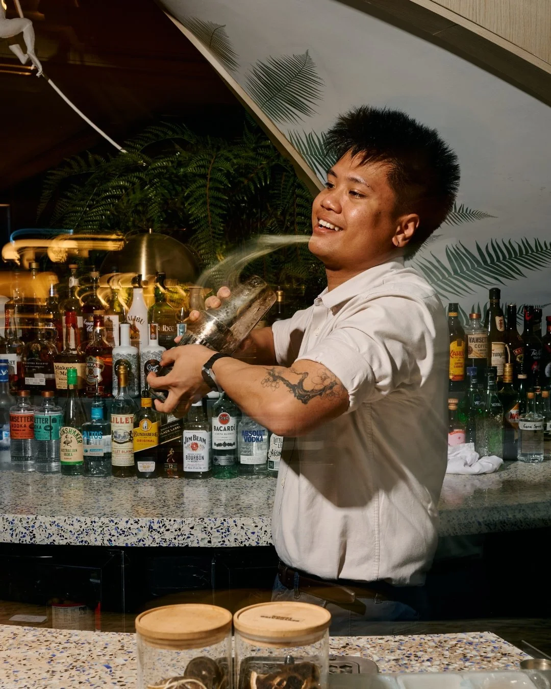 A bartender with short black spiked hair and tattoos on his arm, smiling as he pours a drink at a bar with numerous liquor bottles. There are tropical leaf posters on the wall behind him.