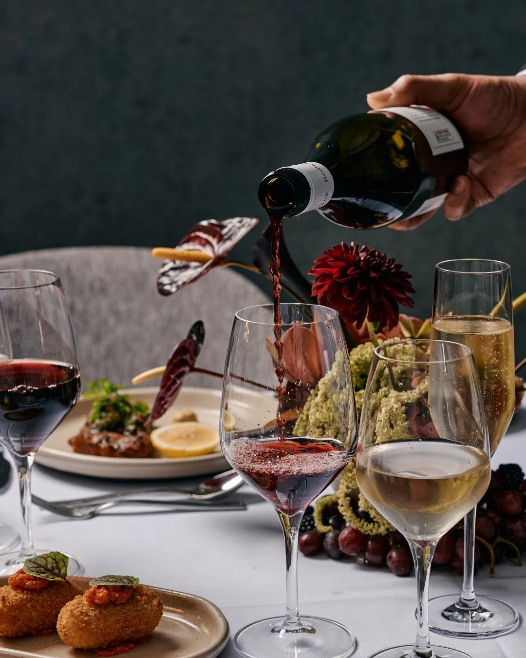 Person pouring red wine into a glass at a table set with glasses of white and sparkling wine, a plate of appetizers, and floral decorations.