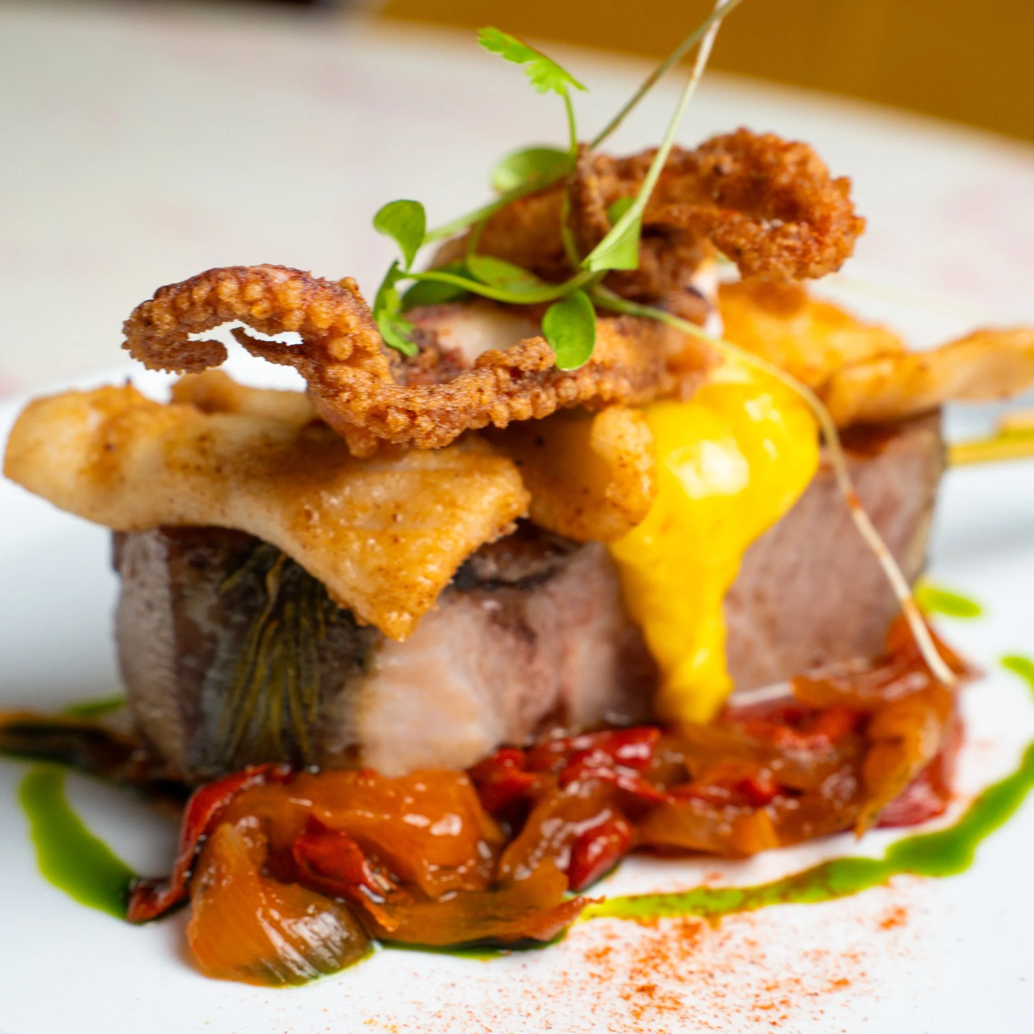 A plated gourmet dish featuring a cooked fish fillet topped with fried calamari, a poached egg with runny yolk, garnished with microgreens, surrounded by roasted peppers, green sauce, and a spice powder.