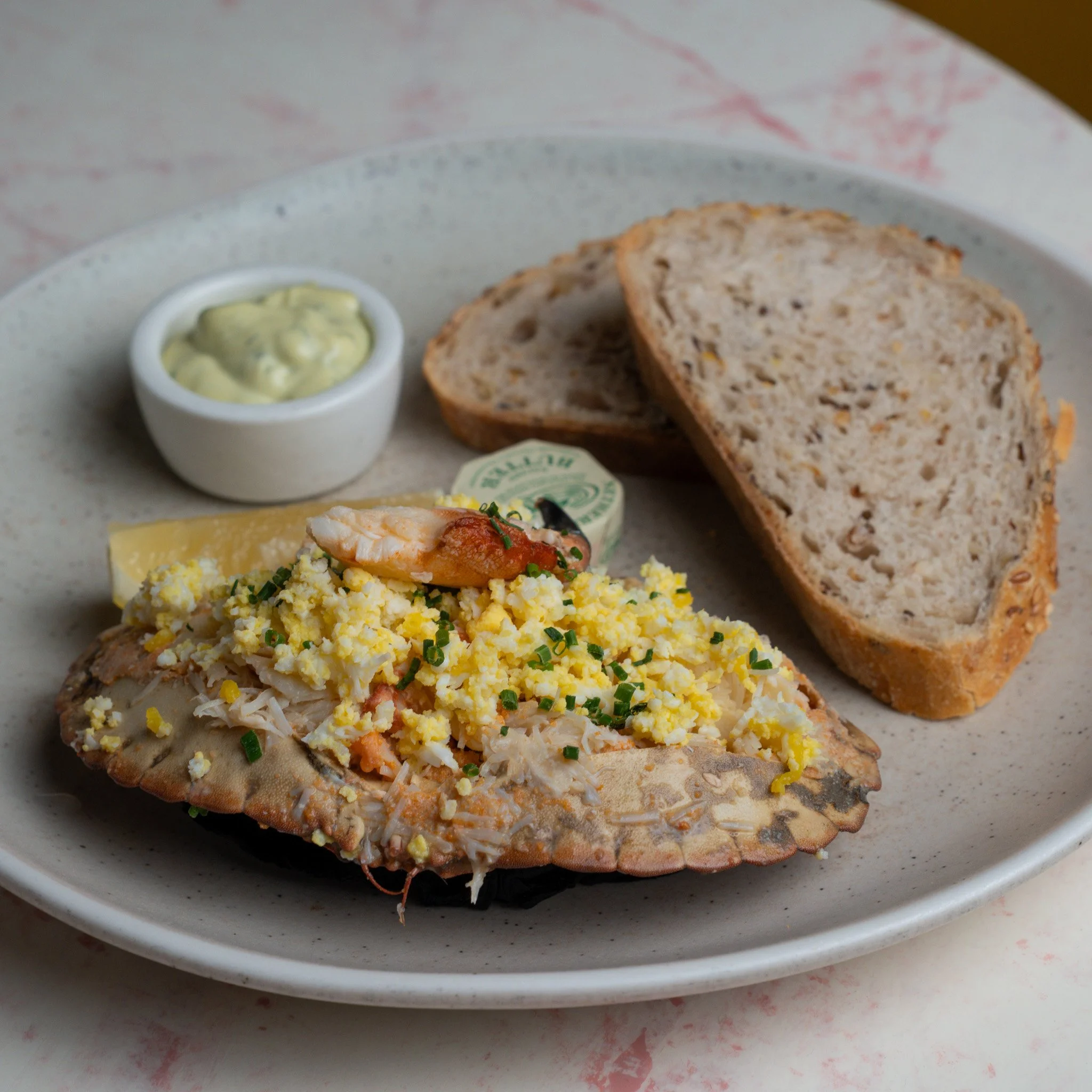 A plate with a fish fillet topped with chopped boiled eggs, a shrimp, a lemon wedge, a small dish of sauce, and two slices of multigrain bread.