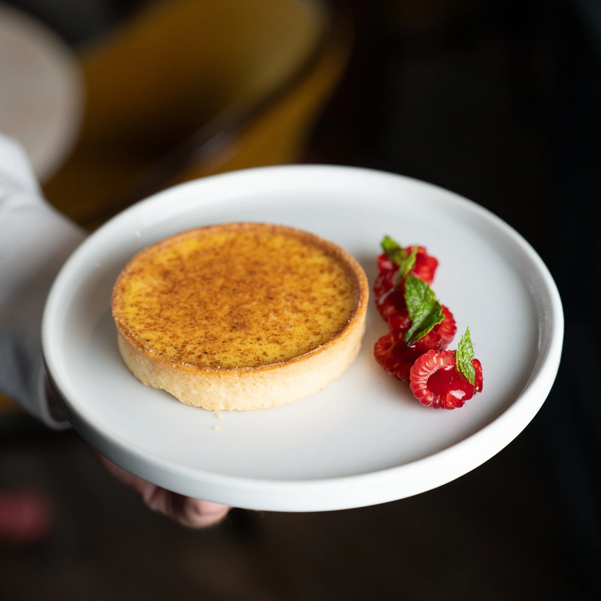 A baked cheesecake with a browned top, served on a white plate with fresh raspberries and mint leaves.