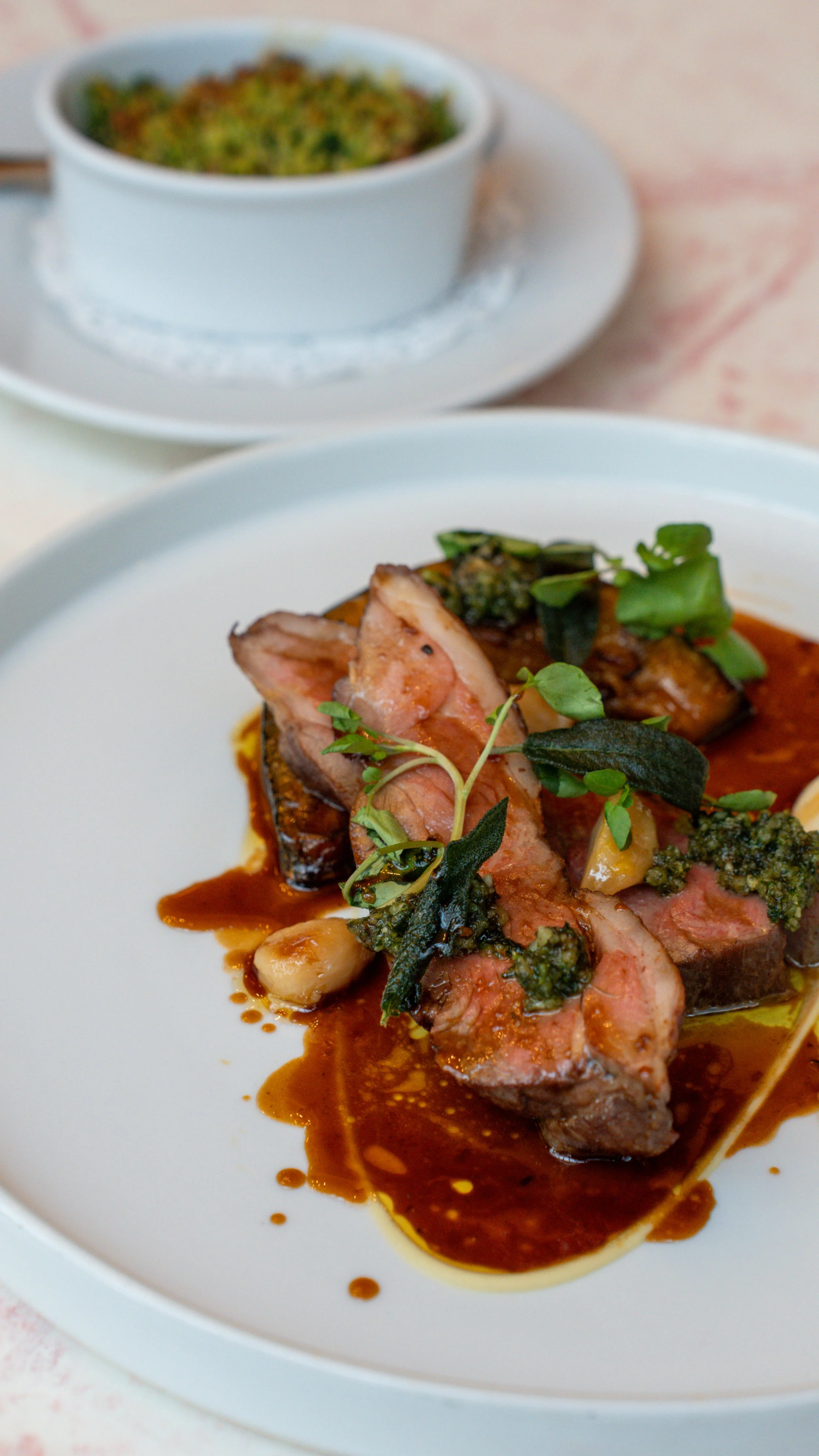 A plate with sliced cooked meat topped with green herbs, sauces, and garnished with small herbs, with a small bowl of baked or casserole dish in the background.