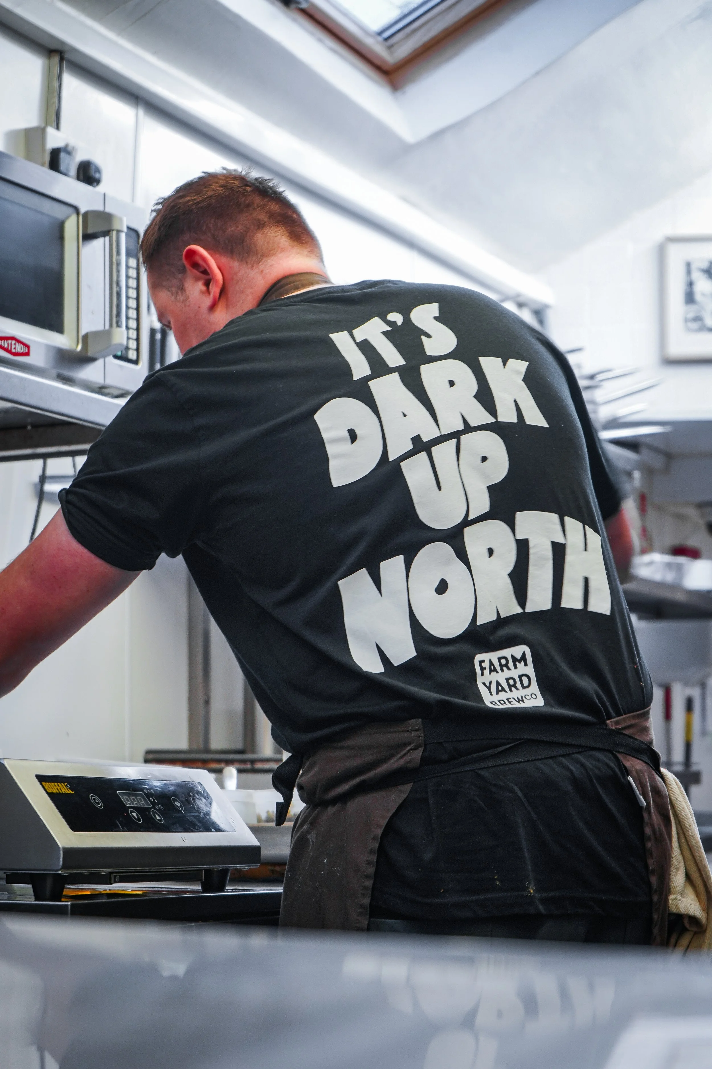 A person in a black T-shirt with the text 'It's Dark Up North' and 'Farm Yard Brewery' on the back, working in a kitchen or food prep area.