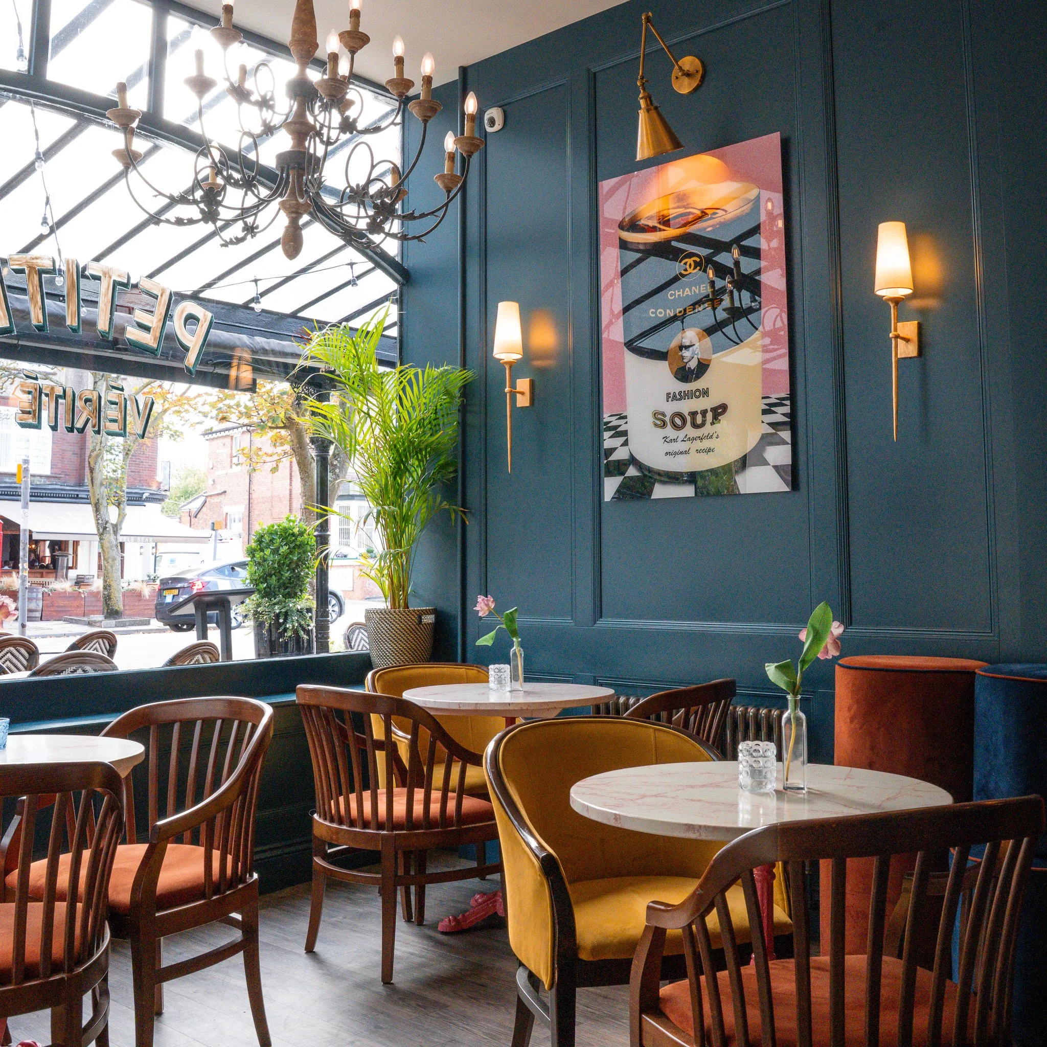 Interior of a cafe with blue walls, framed poster, chandelier, wall lamps, potted plants, and tables with chairs and vases