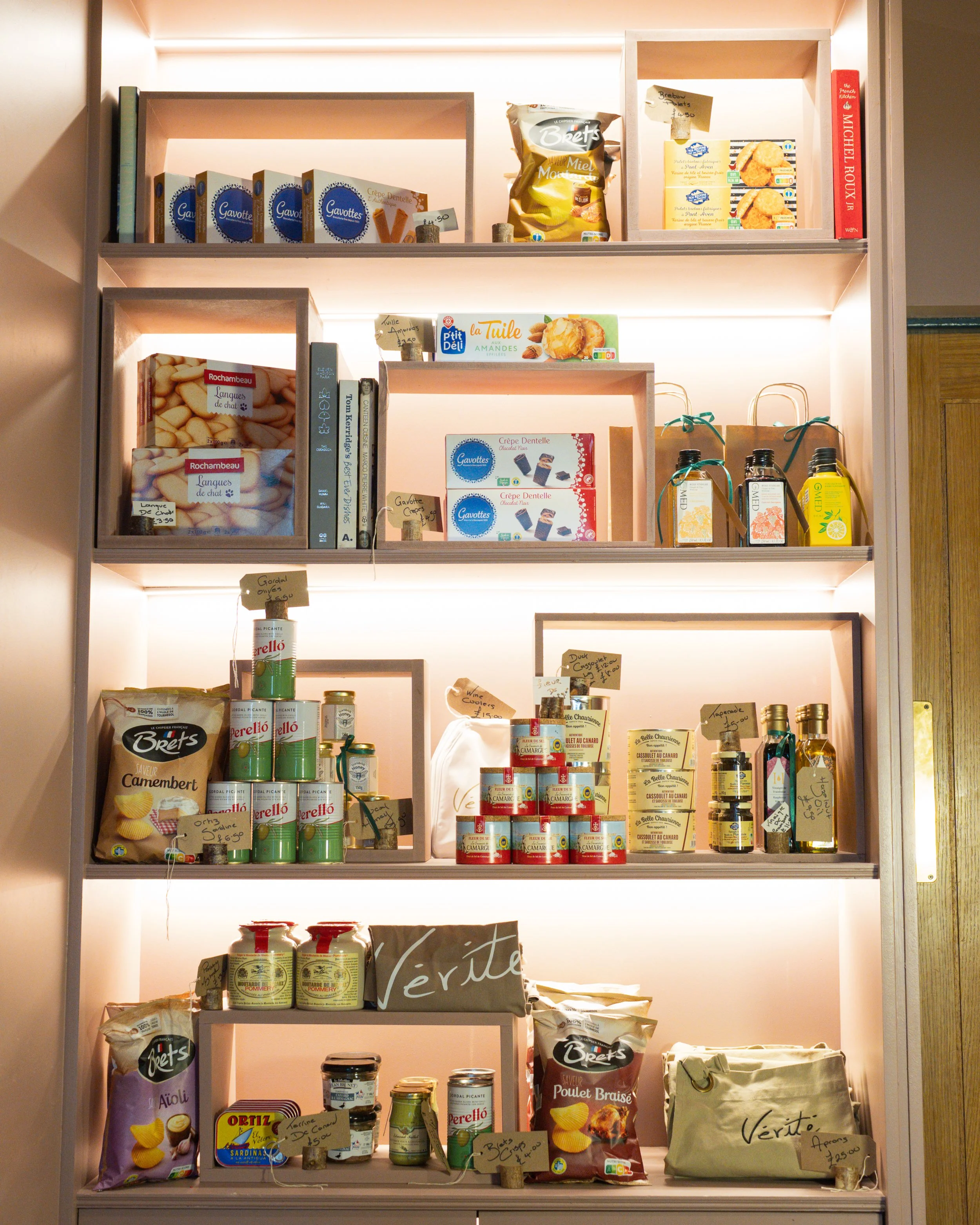 Well-stocked wooden shelves with various packaged food products, including cookies, canned goods, and snacks, with small handwritten price tags attached.