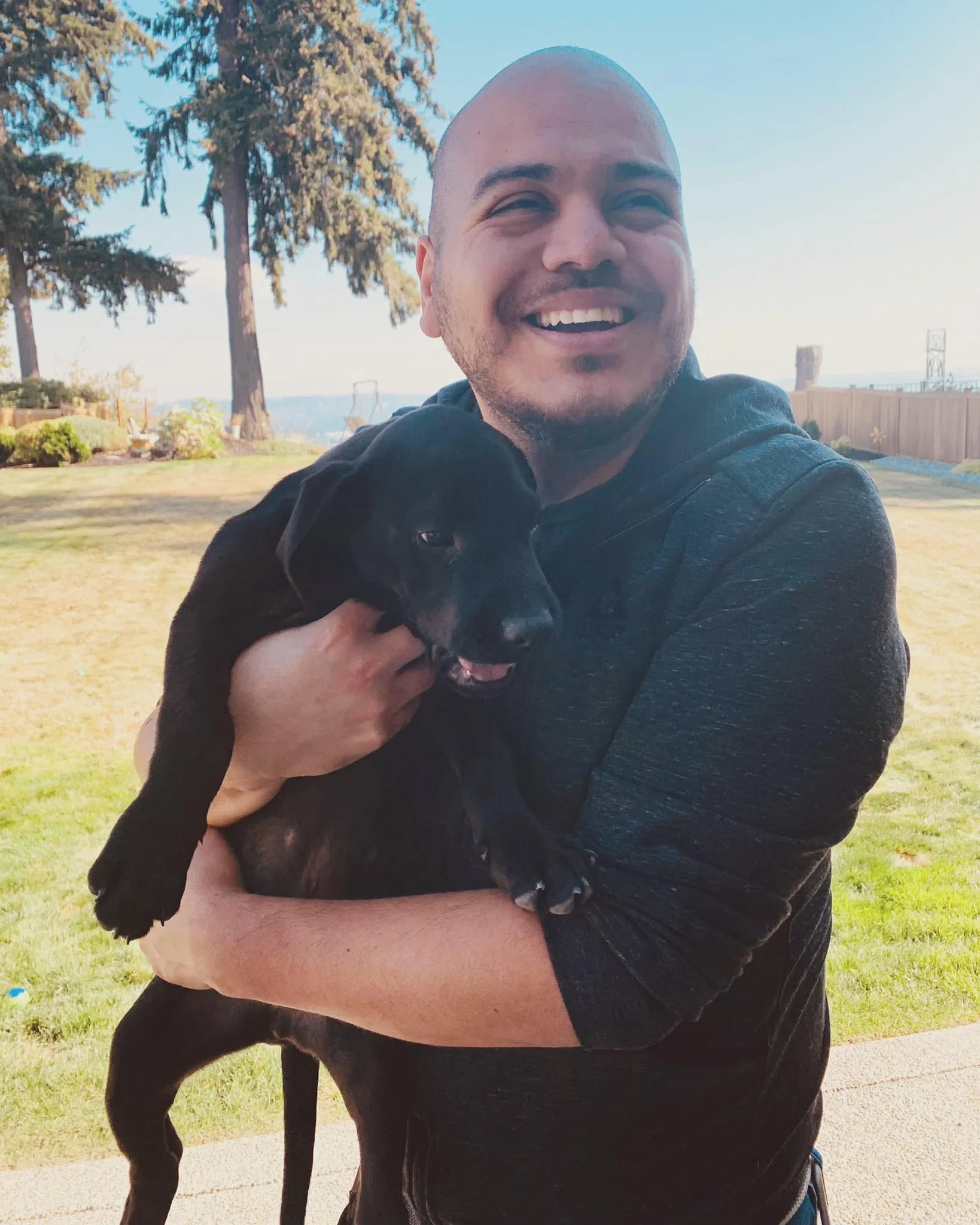 Ollie holding a black puppy outdoors on a sunny day, with trees and a wooden fence in the background.