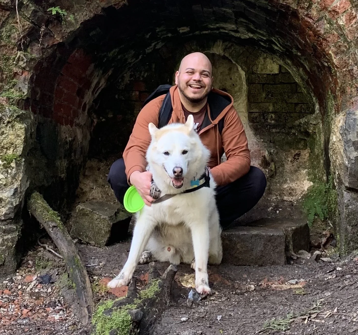 Ollie smiling and squatting behind his white husky dog, Dante, inside a small brick tunnel surrounded by mossy rocks and greenery.
