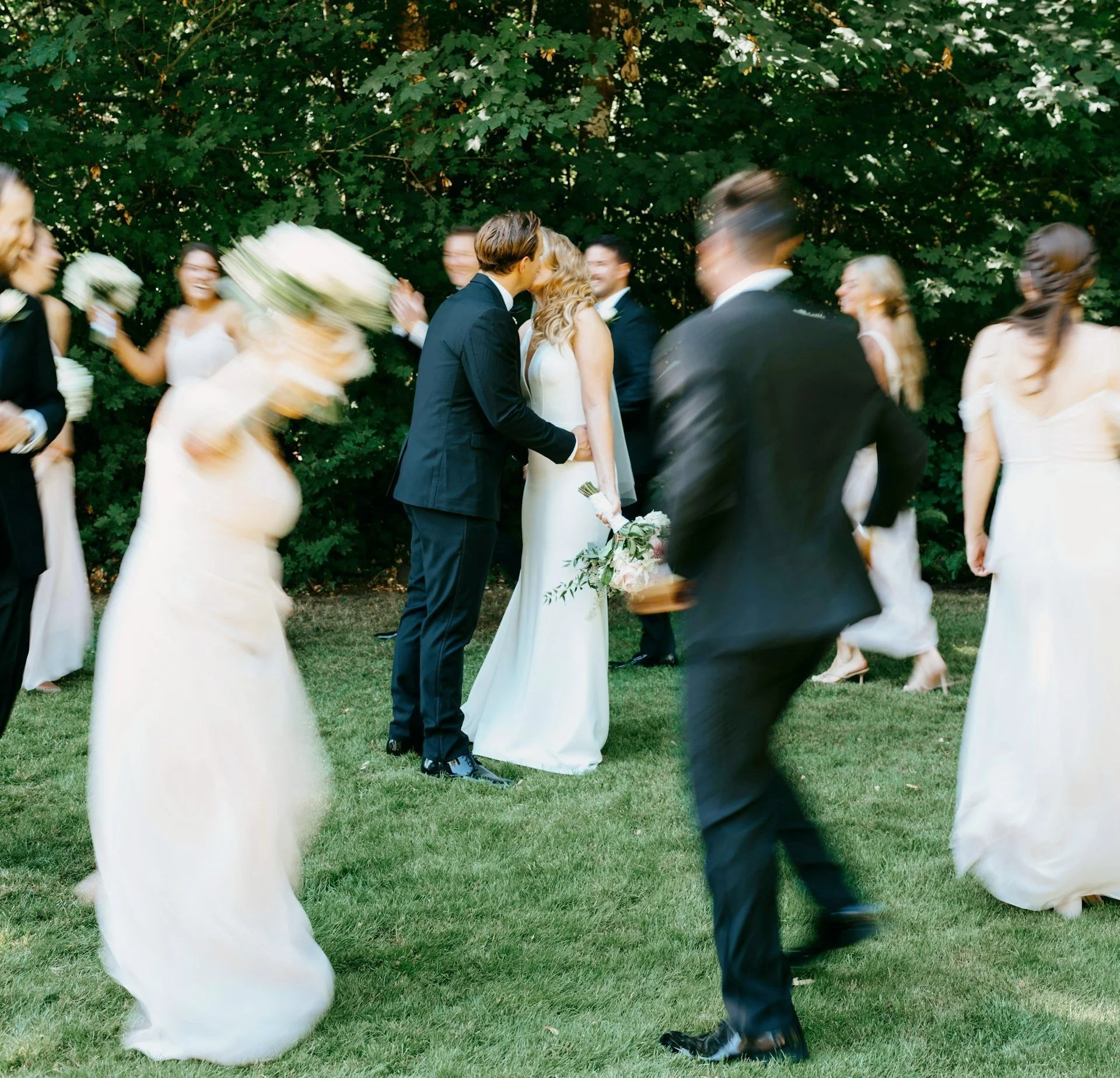 A wedding couple kissing outdoors surrounded by bridesmaids and groomsmen, with some people blurred due to movement.