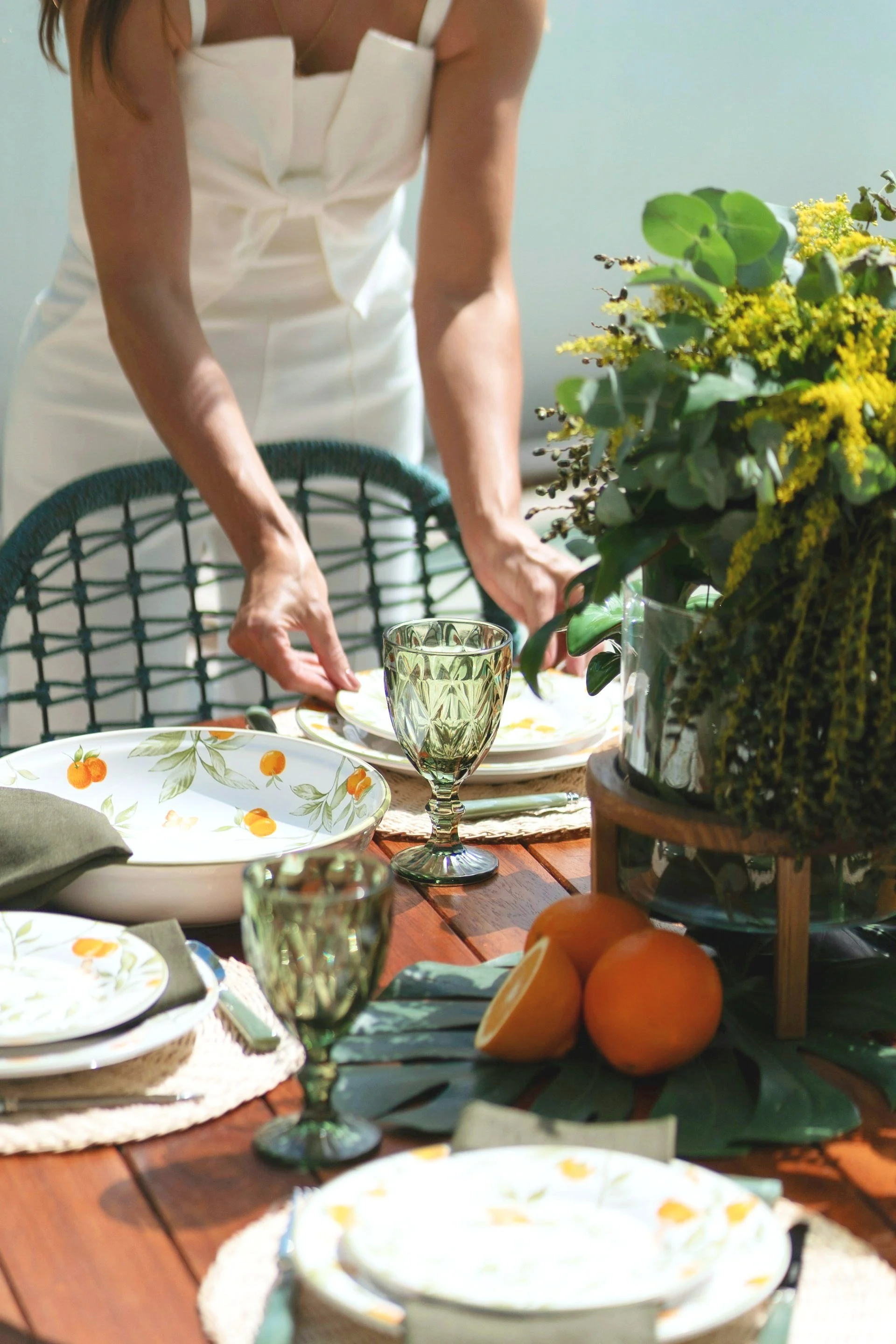 A person setting a table with floral-patterned dishes, green glassware, and fresh oranges, near a large bouquet of yellow and green flowers and leafy decorations.