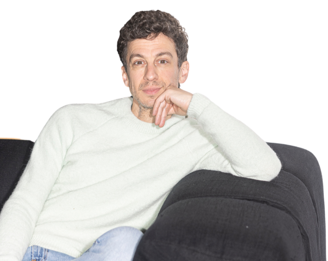 Ein Mann sitzt entspannt auf einem schwarzen Sofa, trägt ein hellgraues Pullover und hat braune, lockige Haare.