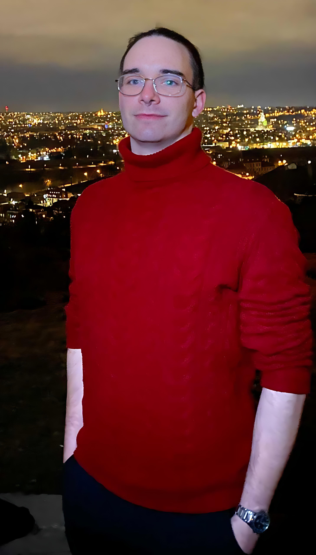 Man with glasses wearing a red turtleneck, standing outside at night with a lit cityscape of Prague in the background.