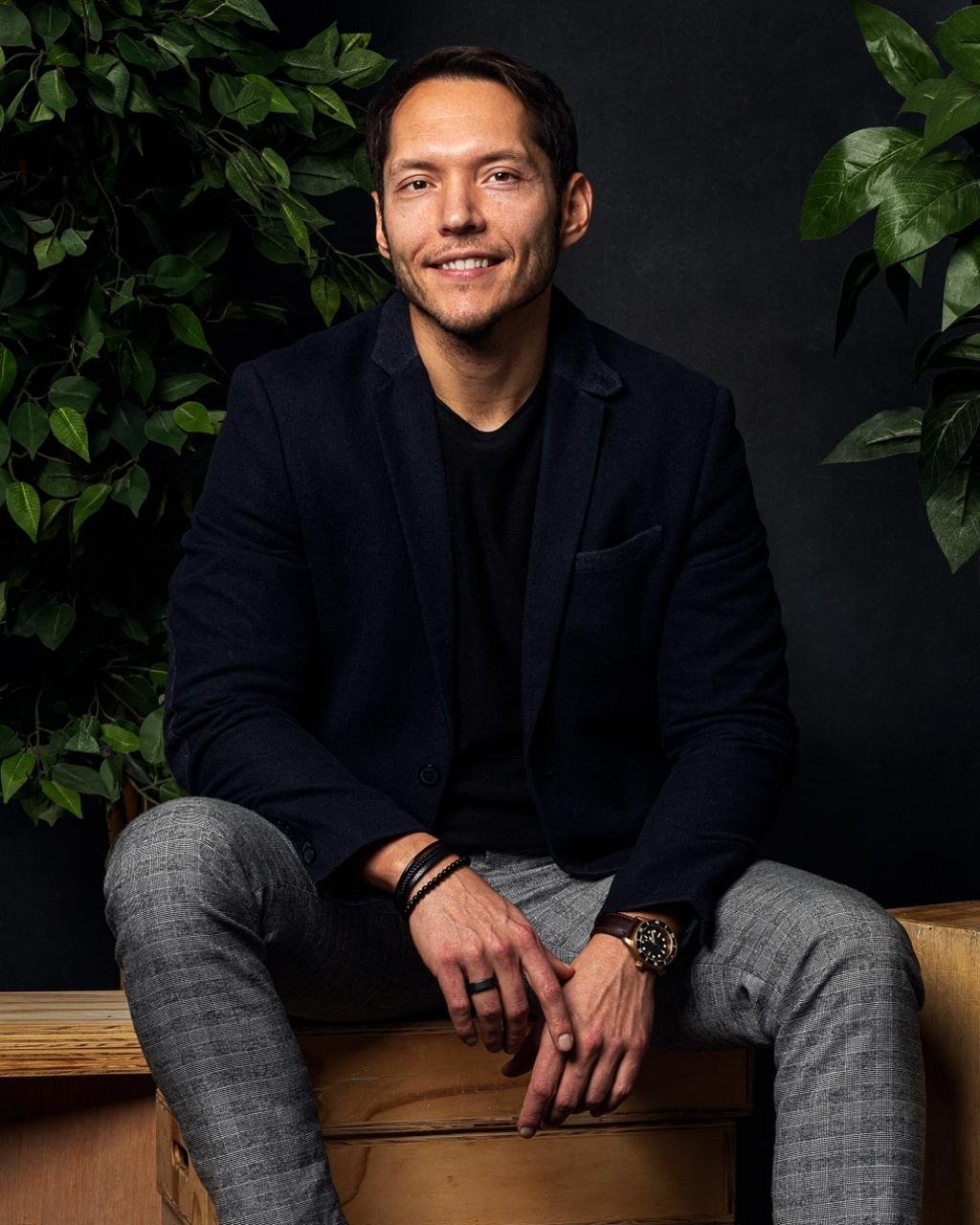 A man with dark hair and a light beard, dressed in a black shirt and a dark blazer, sitting on a wooden surface against a black background with green leaves, smiling.
