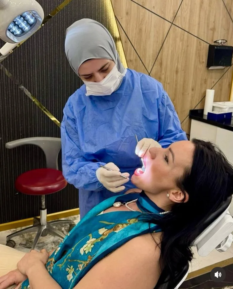 A woman at a dental clinic receiving a dental check-up from a healthcare professional wearing a hijab, face mask, gloves, and blue medical gown, while lying in a dental chair.