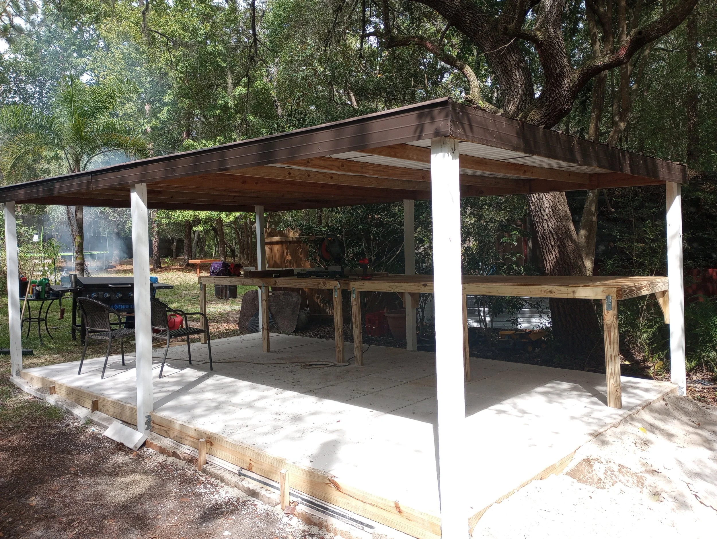 A partially constructed outdoor patio with a roof, wooden beams, and a concrete floor, situated in a backyard with trees and gardening equipment nearby.