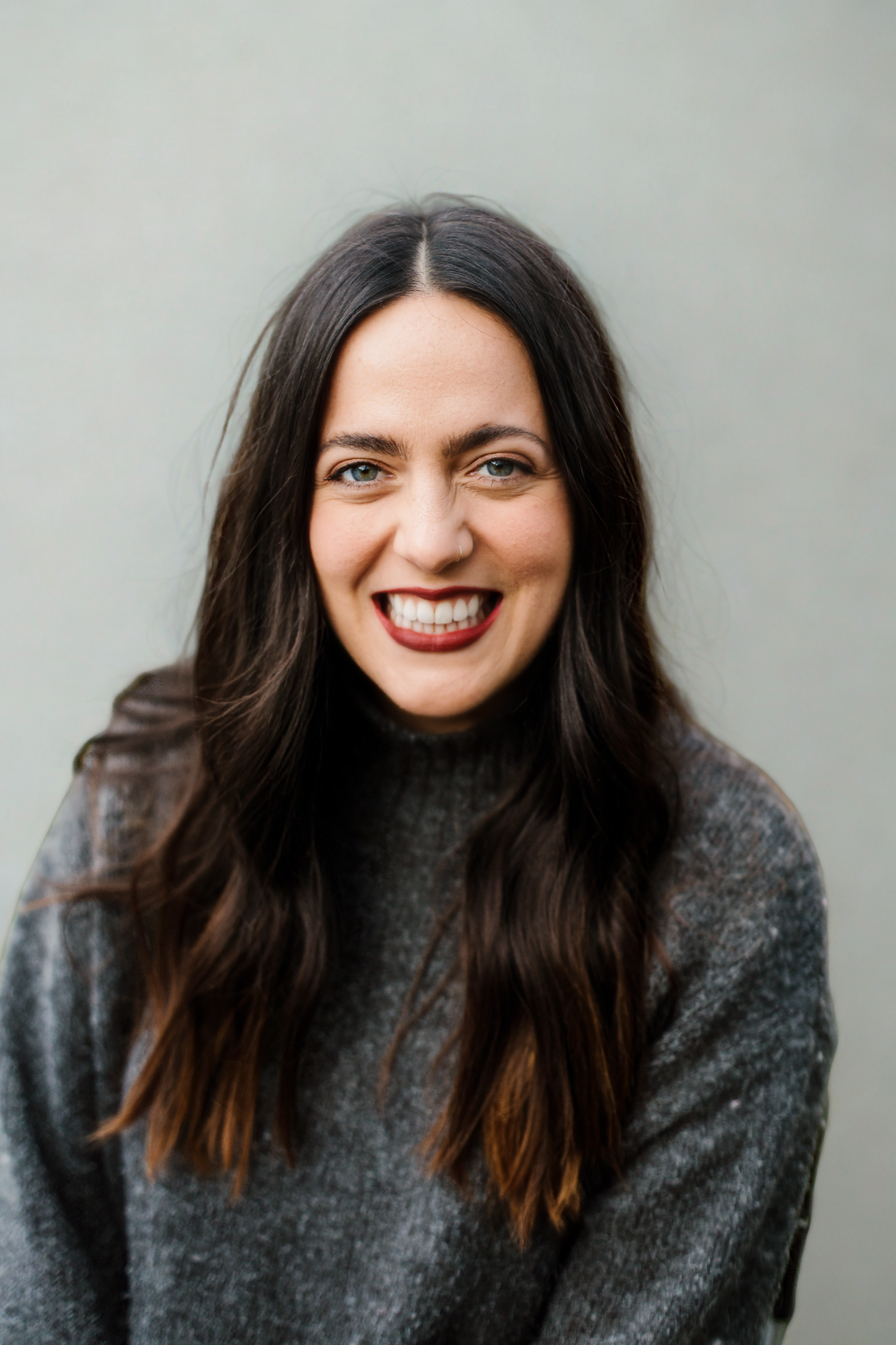 Woman with dark hair and a nose piercing smiling, wearing a gray sweater, against a plain light background.