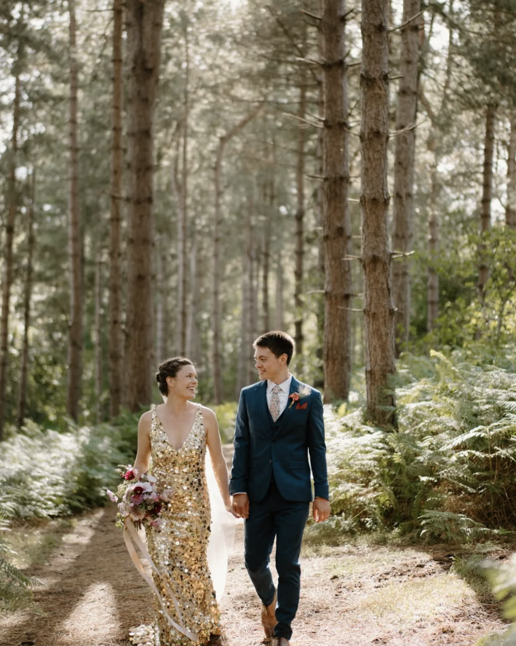 bride in gold sequin dress walks with groom in woodland forest
