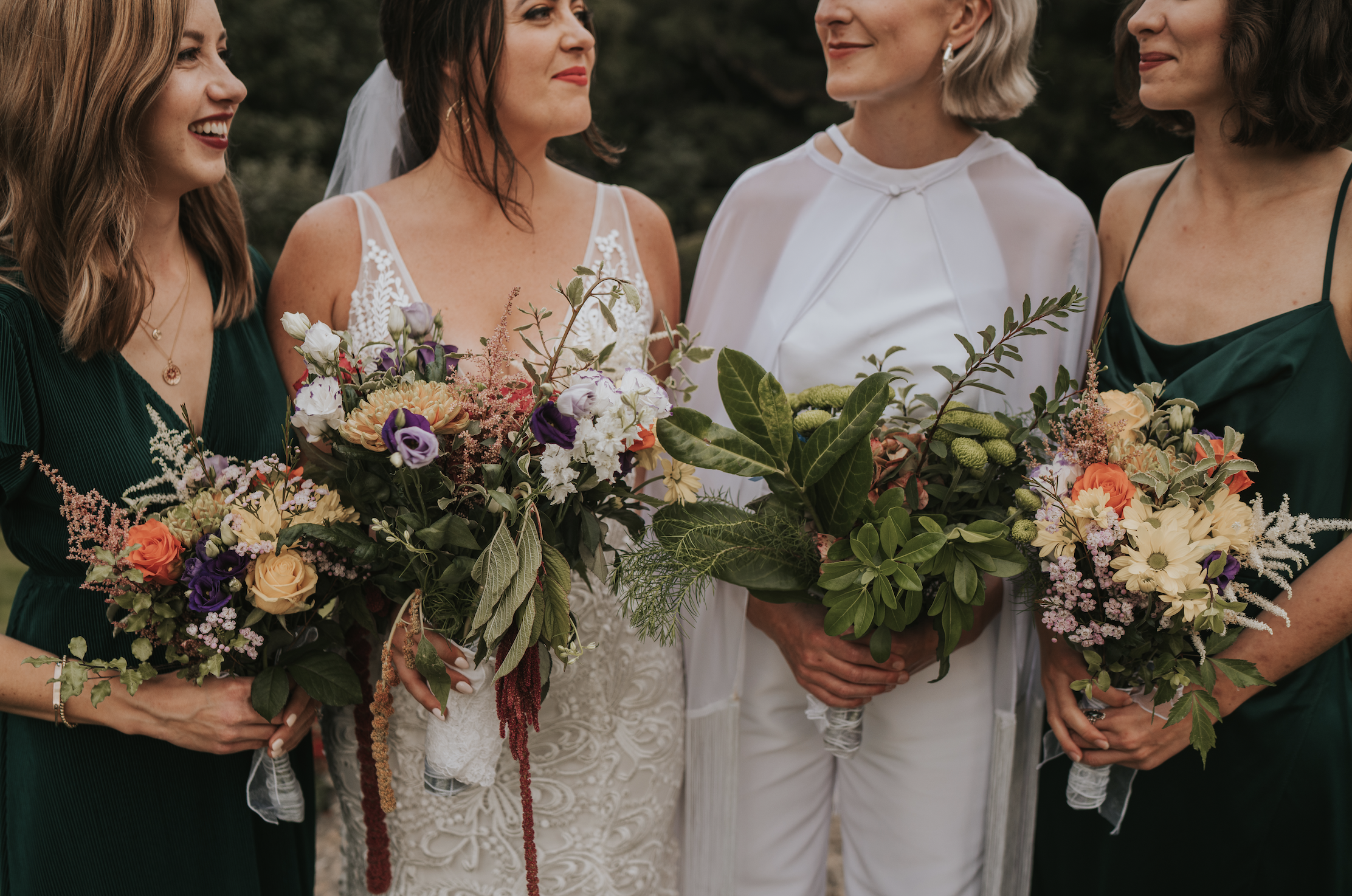 Four women dressed in wedding and bridesmaid attire holding colorful bouquets of flowers, standing outdoors.