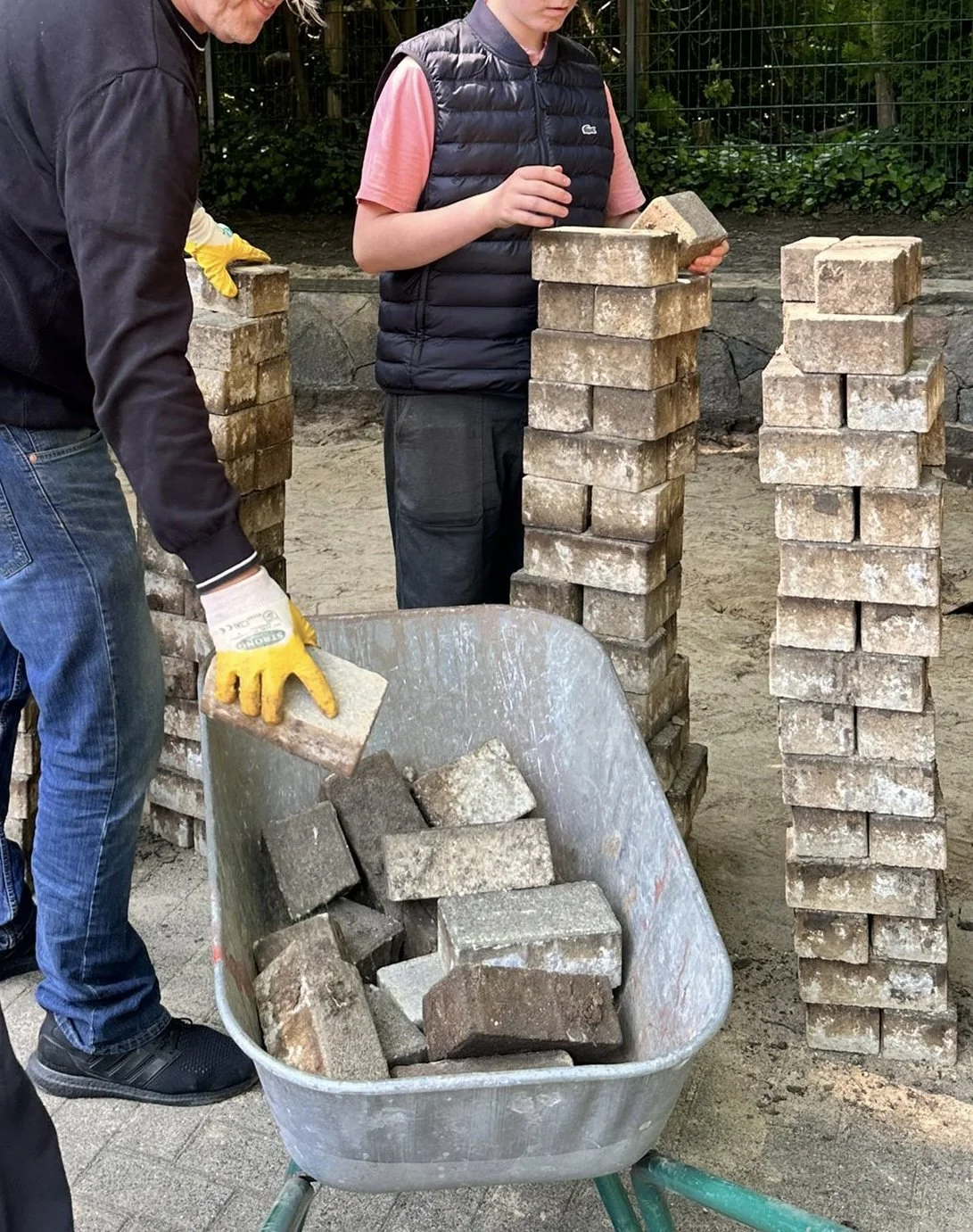 Zwei Personen laden hoch gestapelte Pflastersteine in eine Schubkarre.