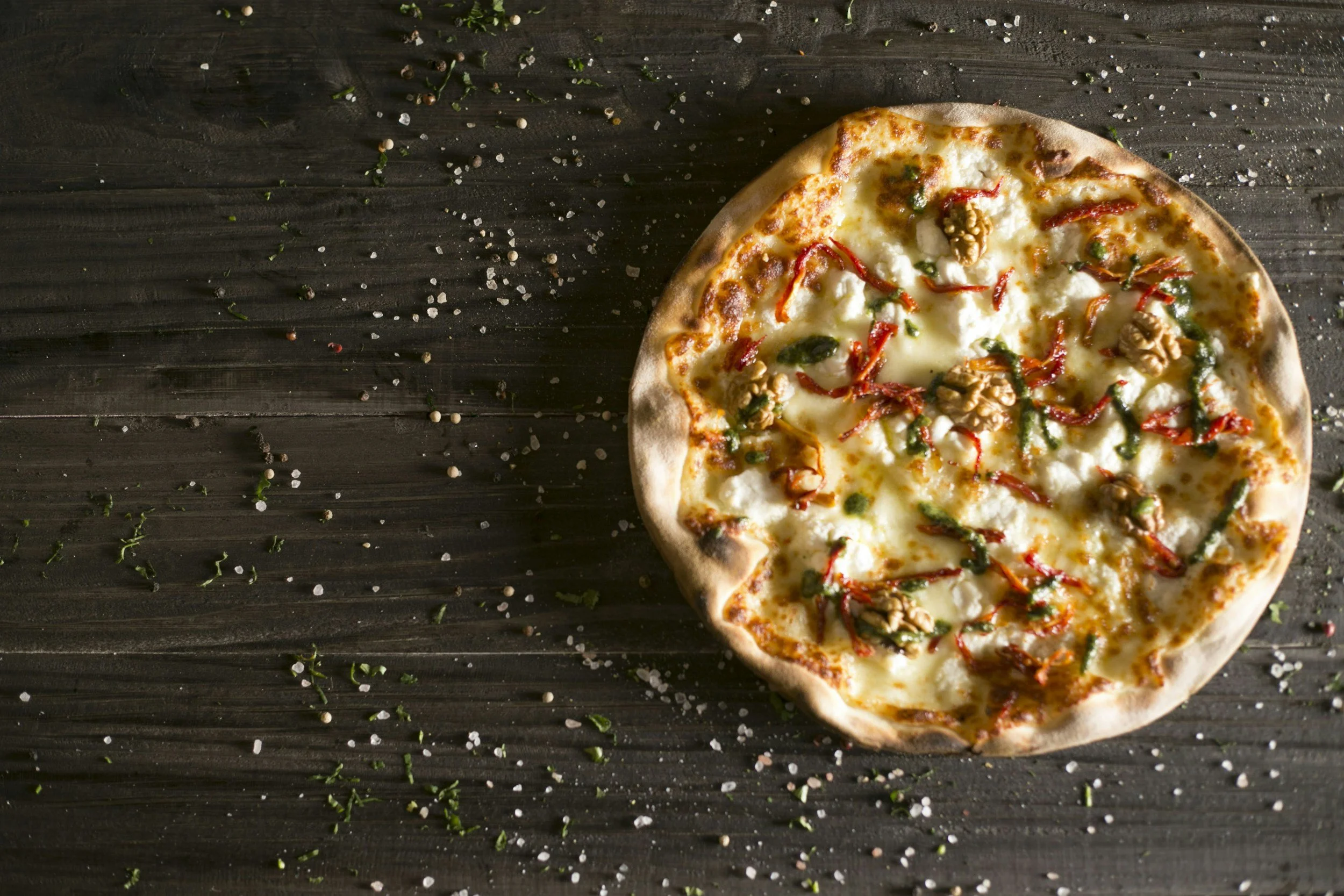 A cheese pizza with red peppers, green herbs, and walnuts on top, placed on a dark wooden surface sprinkled with salt and chopped herbs.