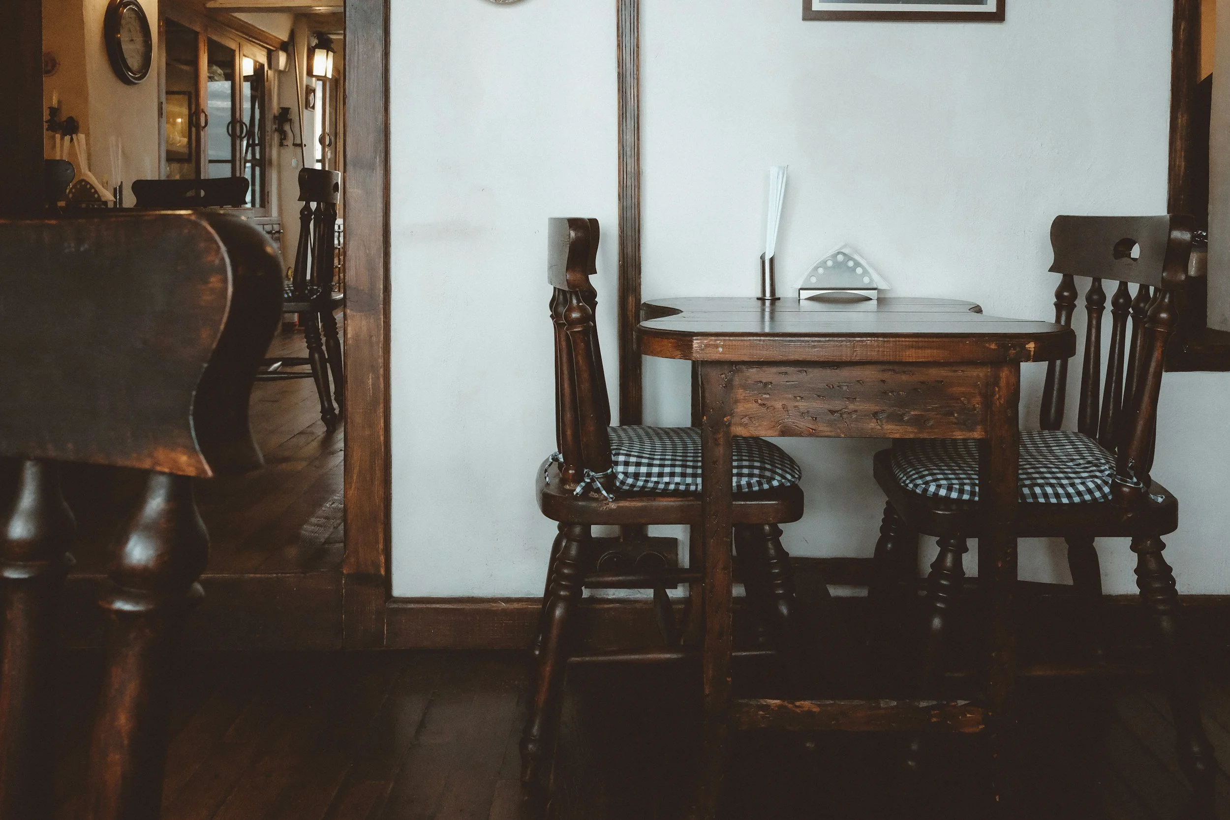 A cozy dining corner with a wooden table and three wooden chairs with checkered cushions, set against a white wall with wooden trim.