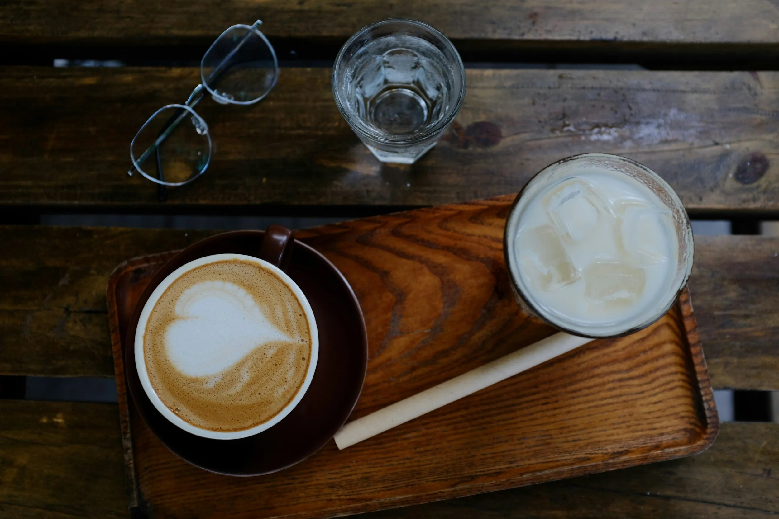 Top-down view of a wooden serving tray with a cappuccino, iced coffee, a glass of water, and sunglasses on a wooden table.