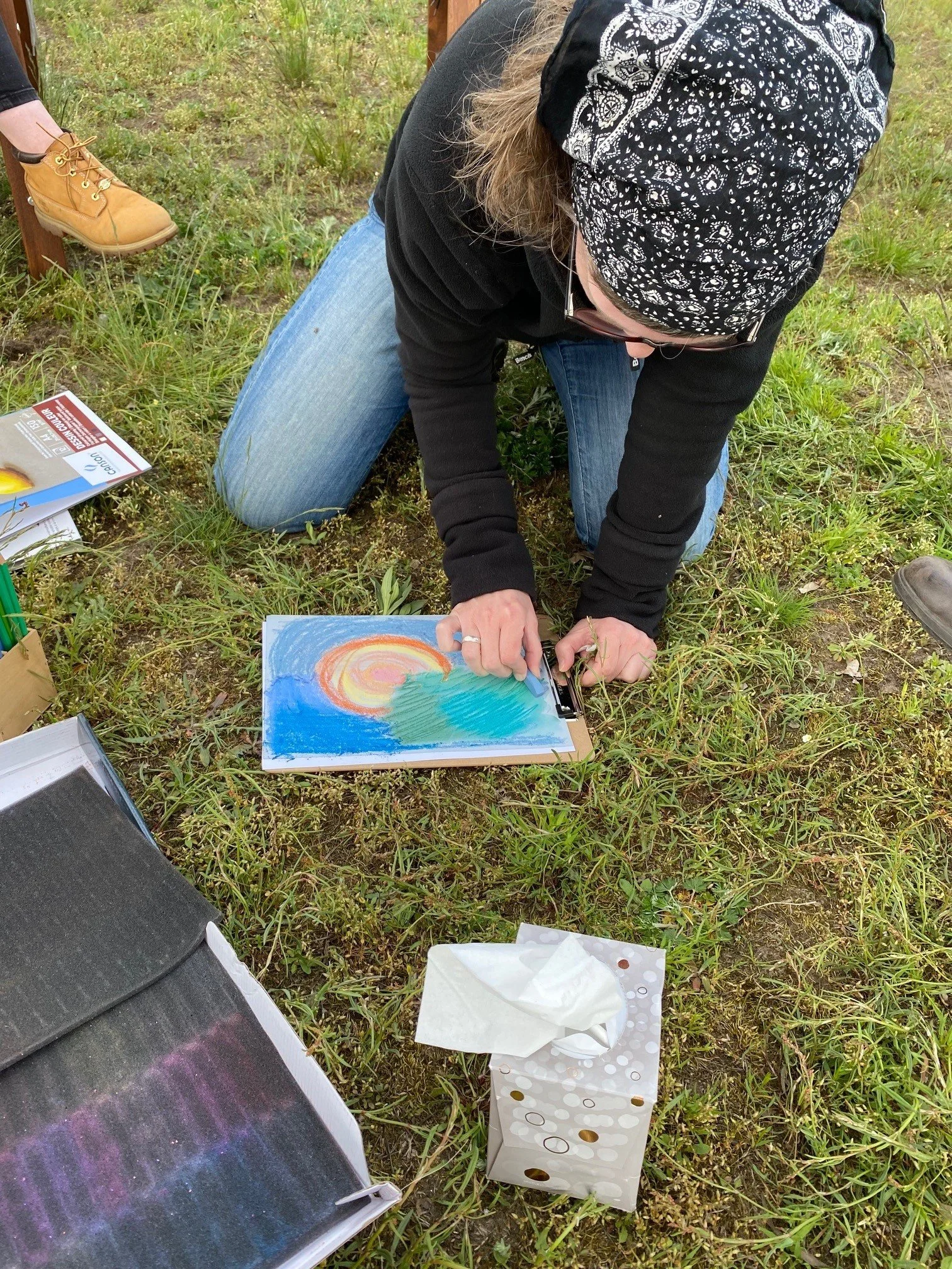 Person beim Malen im Freien, die auf dem Boden kniet, mit einem Bild von einem Sonnenuntergang am Wasser. Es gibt auch Klopapier und andere Zeichenmaterialien in der Umgebung.