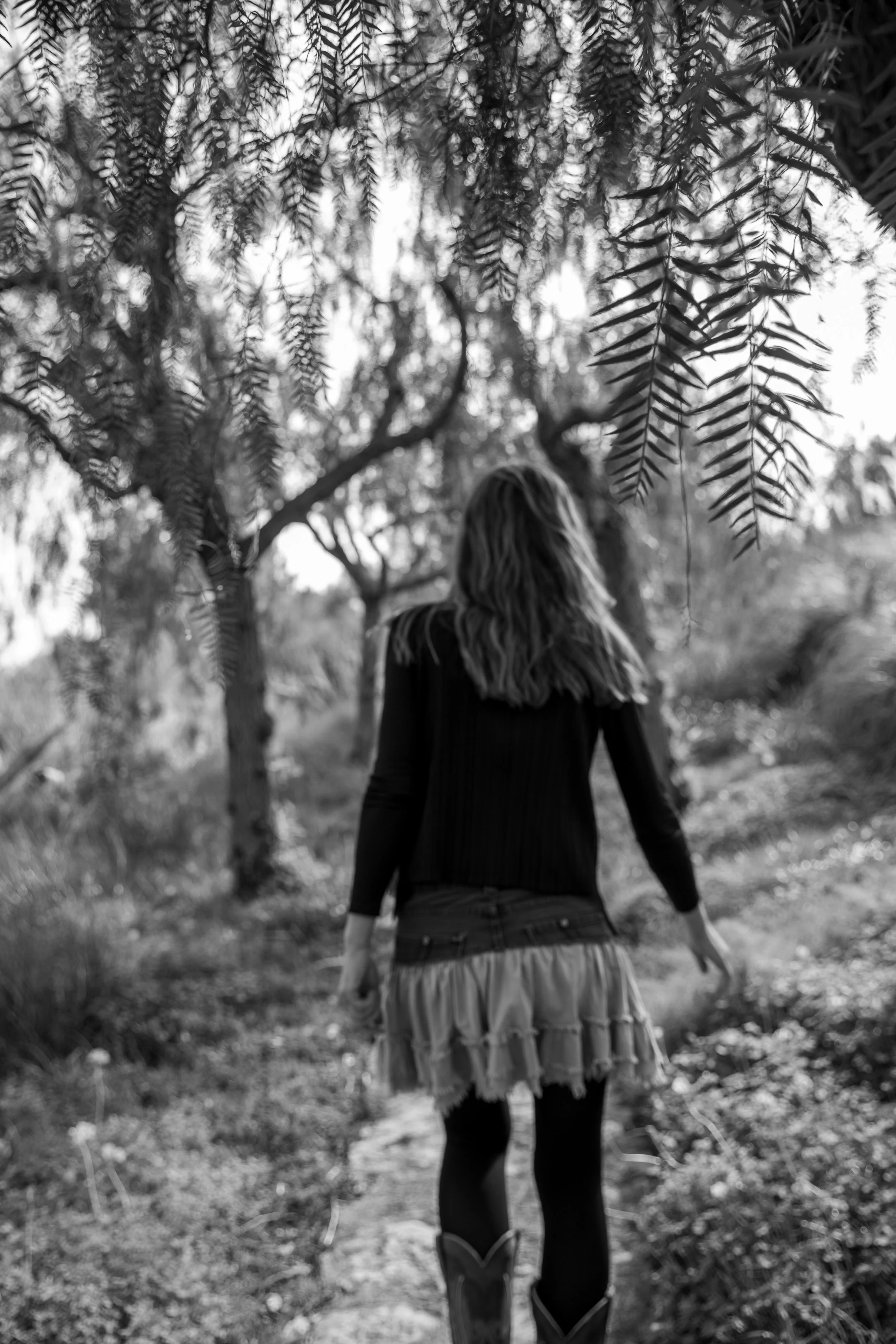 Schwarz-Weiß-Bild einer Frau, die einen Weg in einem Wald oder Park entlanggeht, mit Bäumen und Blättern im Hintergrund, die Oberkörper und Beine sind sichtbar, sie trägt einen Rock, Mütze und Stiefel.