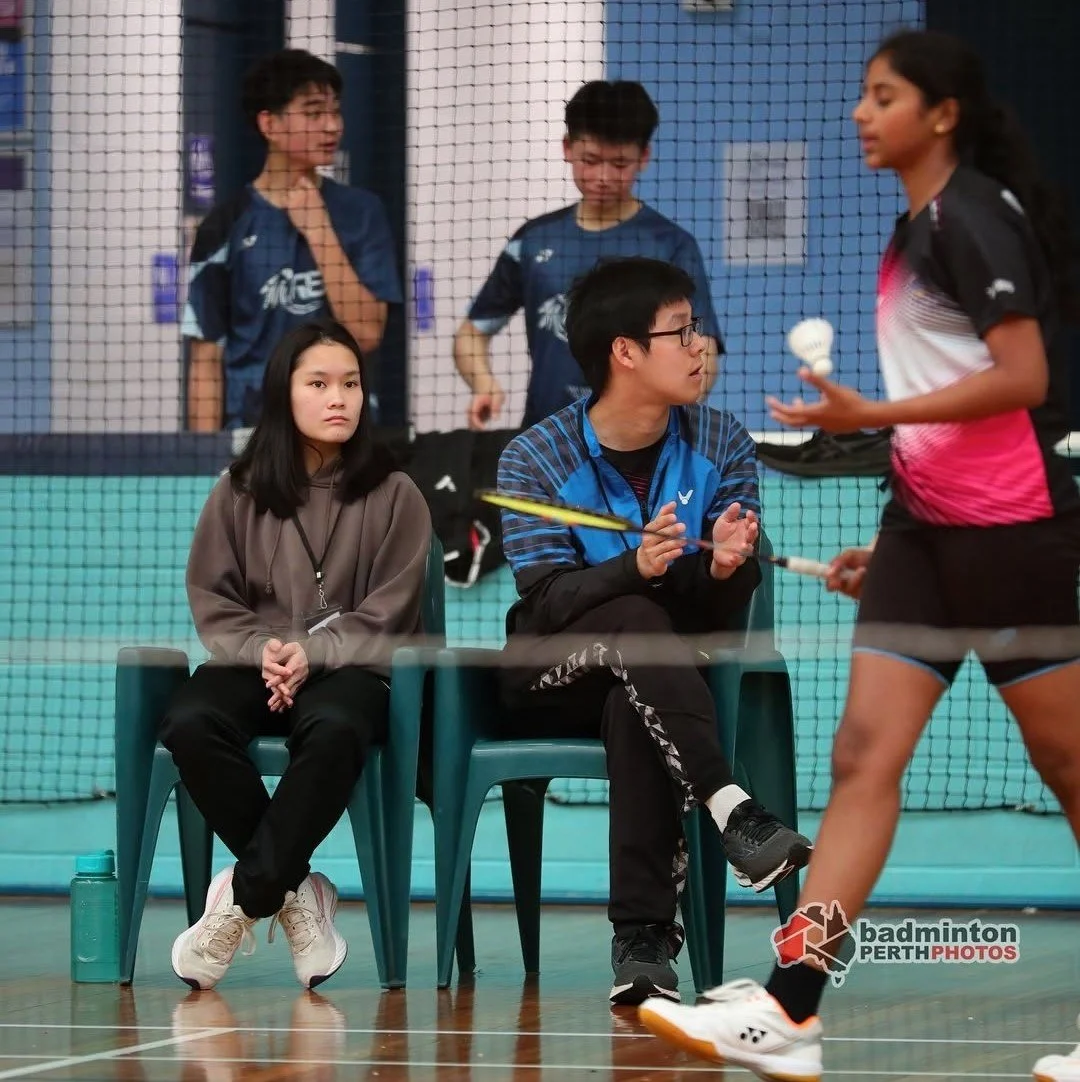 A badminton game with players and coaches in an indoor court. Two women are seated, one is coaching, the other watching. Two young players stand on the court, engaging in a rally.
