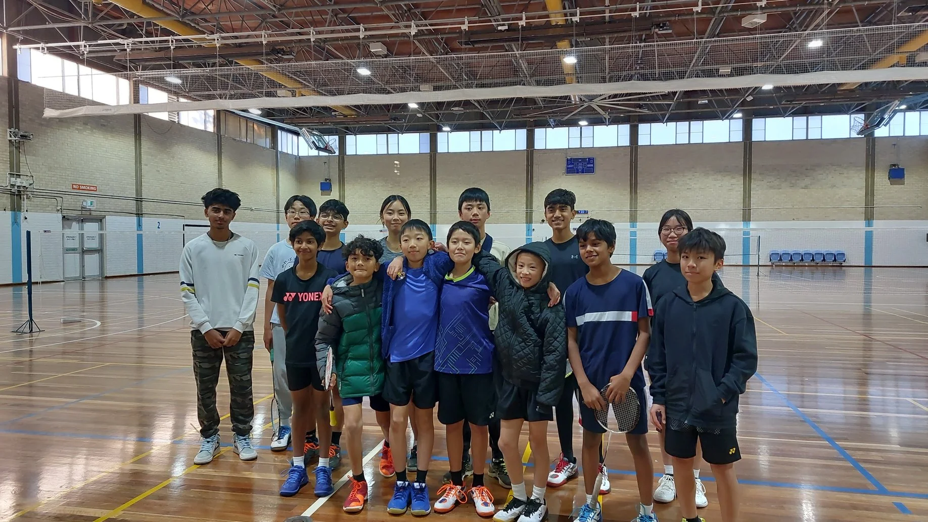 Group of young boys and girls posing on a badminton court inside a gymnasium.