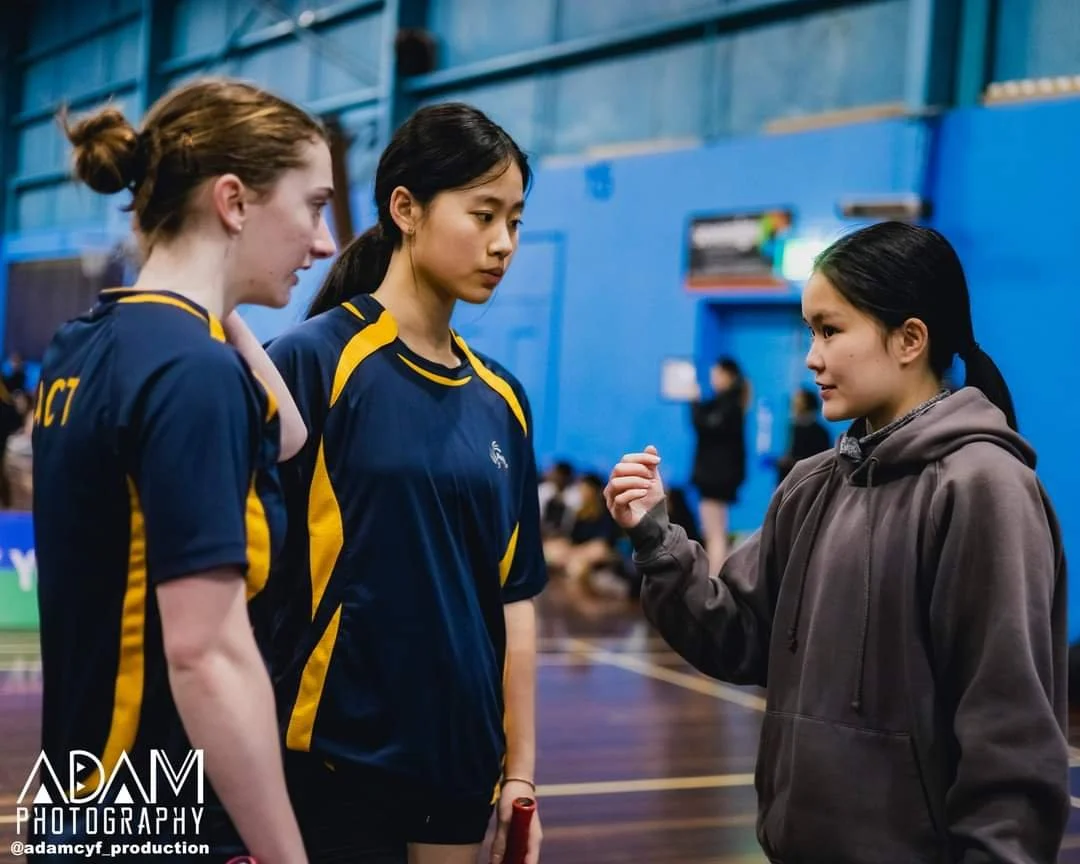 Three young women are having a serious conversation in a gymnasium. Two of them are wearing navy blue sports jerseys with yellow accents, and the other is dressed in a gray hoodie. The woman in the hoodie is gesturing with her right hand, while the two women in jerseys listen attentively.