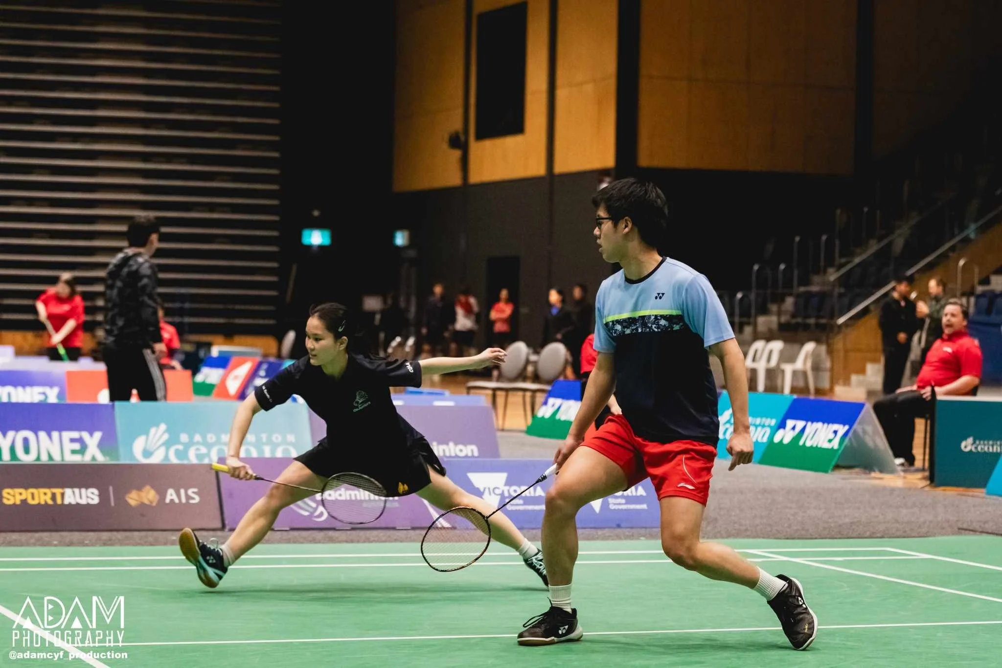 Two badminton players in action on a court indoors, with one female reaching for a shot while the male player stands nearby. Several spectators and officials are visible in the background, with banners and advertisements along the sides of the court.