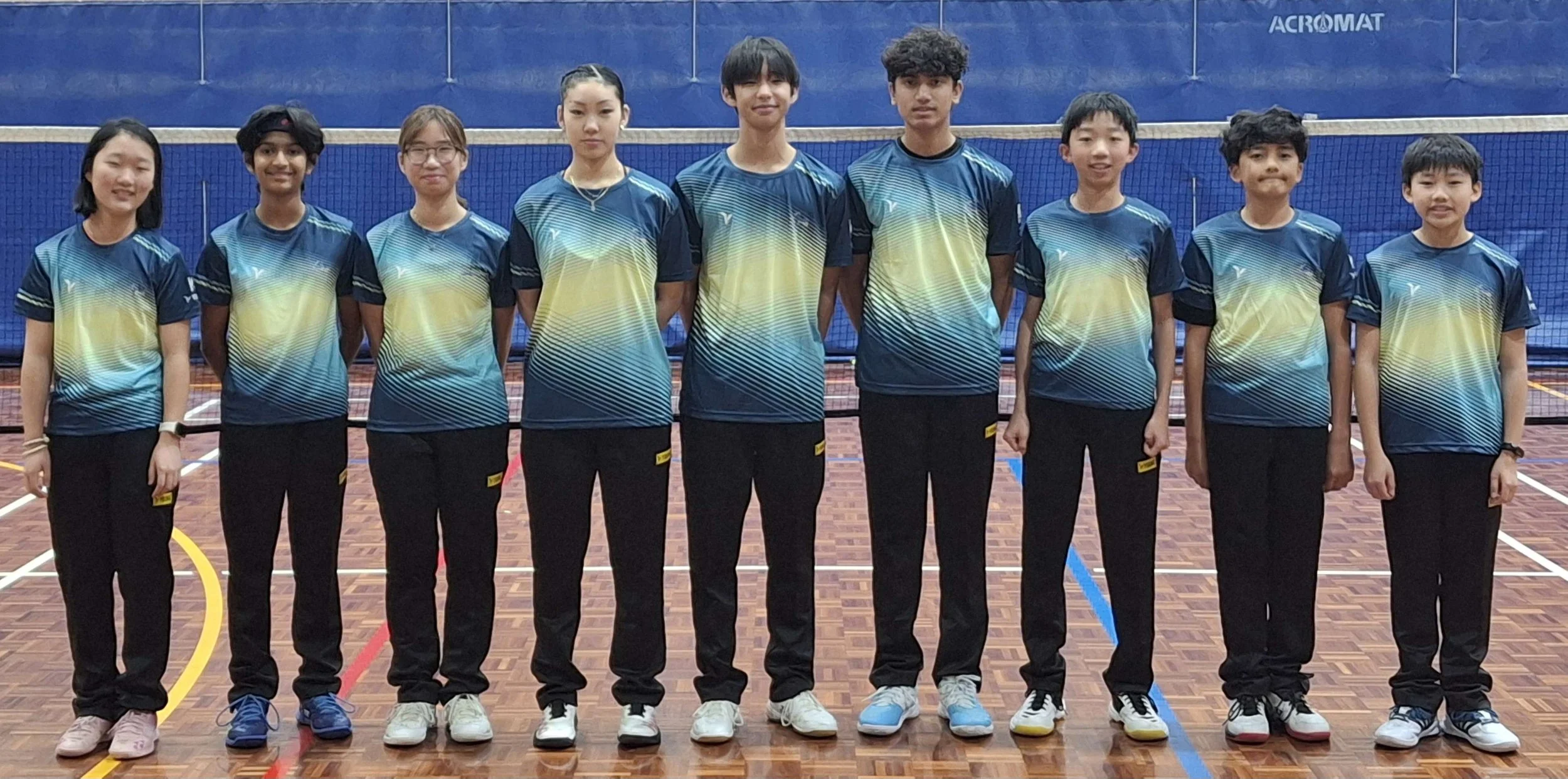 Group of ten young athletes standing in a row on an indoor sports court with blue backdrop. They are wearing matching sports jerseys and black pants, smiling and facing the camera.